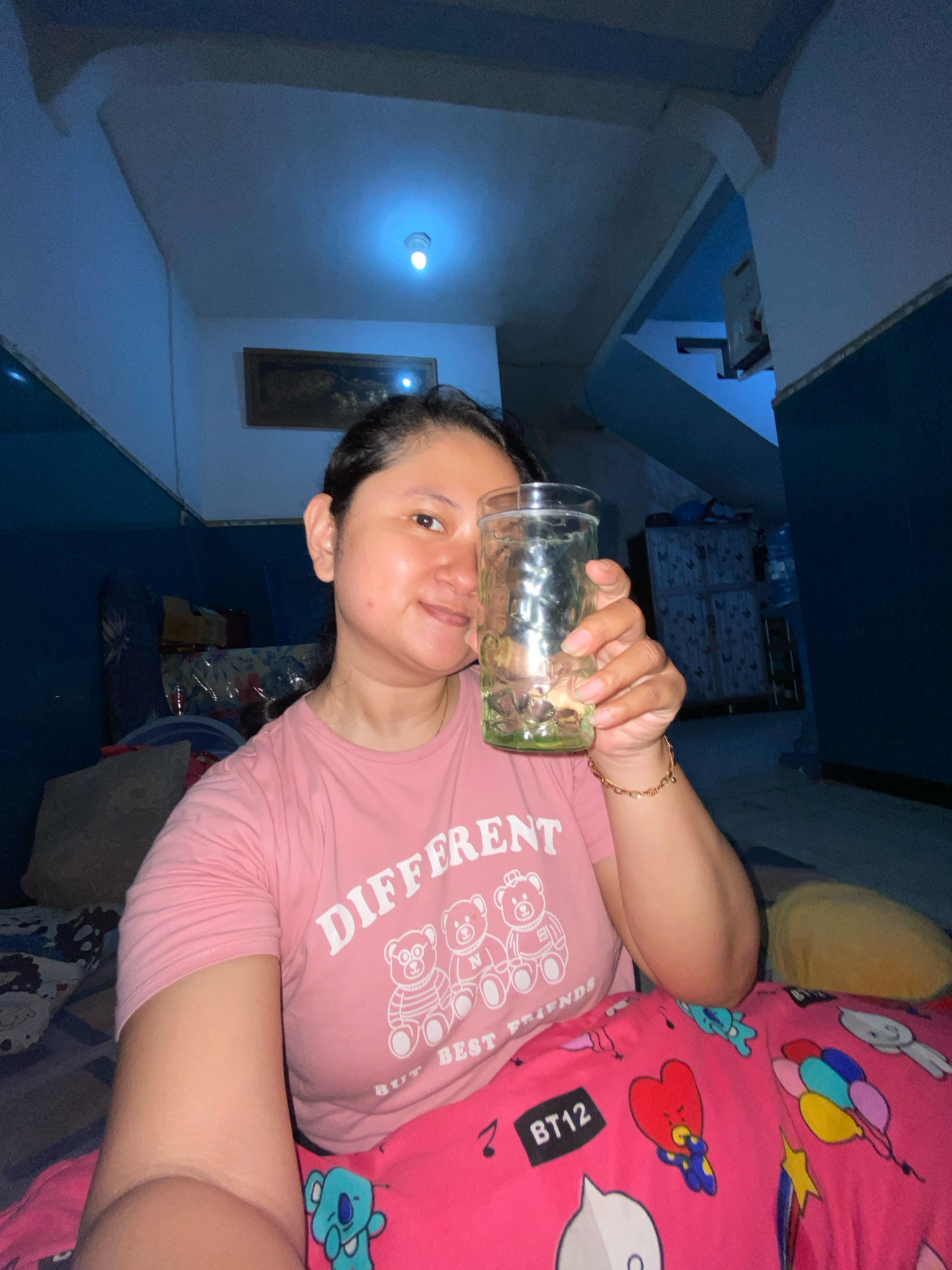 A medium shot captures a woman sitting on a bed in Gelam, Indonesia, holding a glass of water. The room is dimly lit, with a ceiling light casting a blueish glow. The woman wears a pink t-shirt with a graphic design of bears and text that reads "DIFFERENT BUT BEST FRIENDS". Her lower body is covered by pink pajama bottoms adorned with cartoon characters and the text "BT12". The background reveals a dark room with blue walls and a staircase.