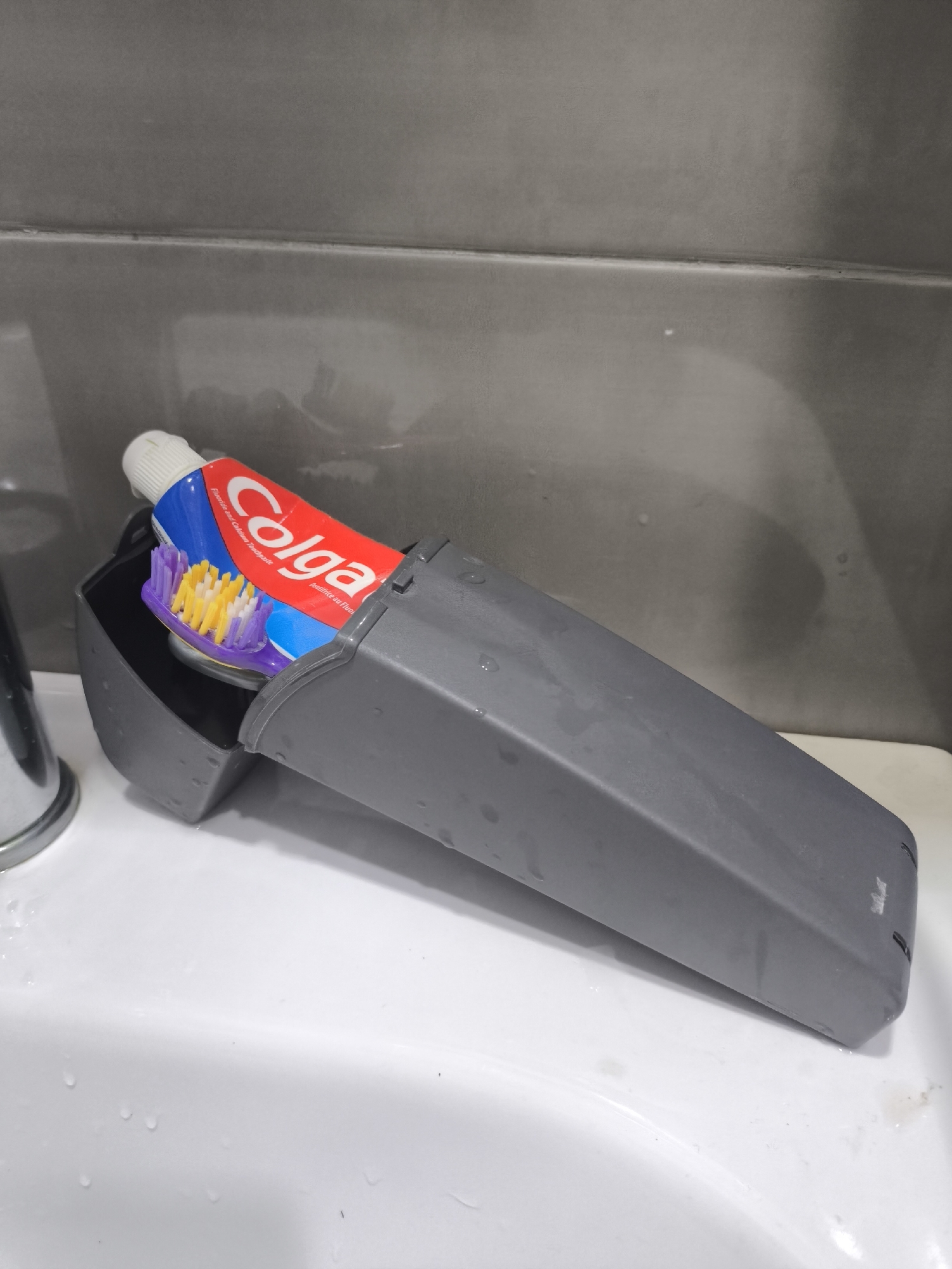 In Batumi, Georgia, a grey plastic holder is positioned on the edge of a white sink. Inside the holder, a tube of Colgate toothpaste is placed at an angle, with its cap off. A purple and yellow toothbrush is nestled alongside the toothpaste, with its bristles pointing towards the tube. The background consists of grey tiled walls. Water droplets are visible on the sink surface and the plastic holder.