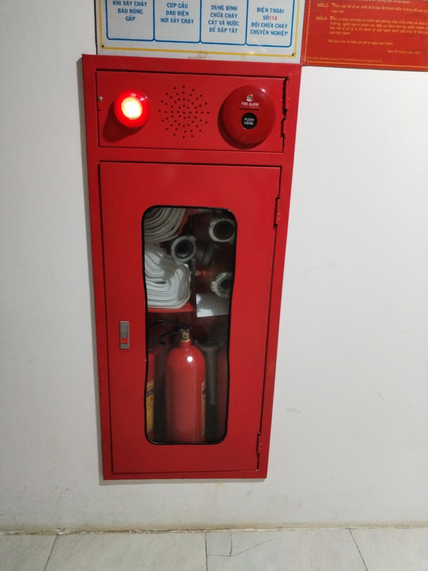 The image captures a fire safety station mounted on a white wall, likely within a building in Bac Nha Trang Ward, Vietnam. The station is bright red and features two main compartments. The upper compartment includes a flashing red light, a speaker grill, and a "FIRE ALARM" button labeled "PUSH HERE". The lower compartment, visible through a transparent window, contains neatly coiled fire hoses and a red fire extinguisher. Above the station is a series of signs in Vietnamese, including instructions for fire emergencies, such as "KHI XÂY CHÁY BÁO ĐỘNG GẤP" (When a fire occurs, alert urgently). A partially visible red sign on the right outlines fire safety regulations and rewards for fire prevention efforts. The overall environment appears to be an interior space with tile flooring.