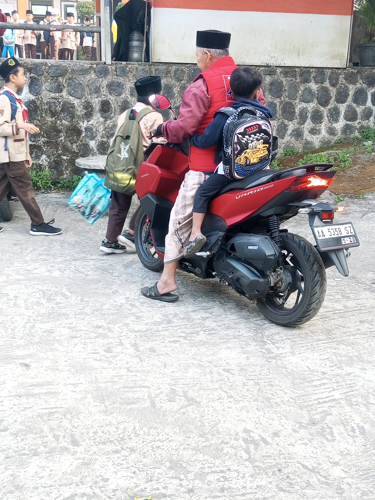 The image depicts a street scene in Krasak, Indonesia, focusing on a red motorcycle with three occupants. An adult male in a red vest and a black peci hat is driving, with a young boy wearing a backpack and a blue jacket sitting behind him. Another young boy in a scout uniform is visible to the left, walking away from the motorcycle. In the background, several other children in scout uniforms are lined up, and a stone wall runs along the edge of a building. The ground is paved with a textured concrete. The overall impression is one of a school dismissal or a gathering of students.