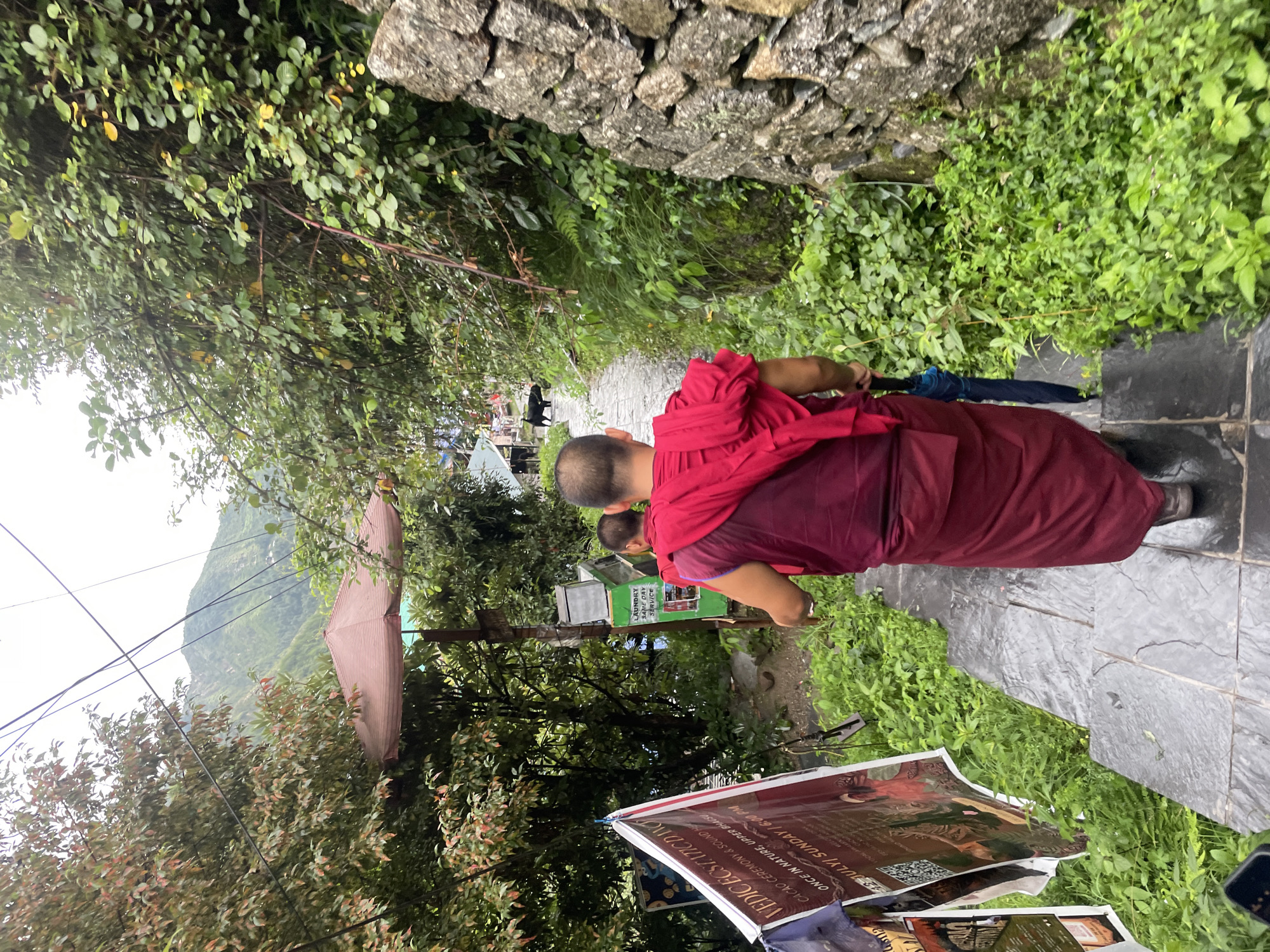 A Buddhist monk in maroon robes walks down a stone staircase, holding a blue umbrella. The staircase is surrounded by lush green vegetation and a stone wall. In the background, a black animal, possibly a yak or cow, walks along a path. There is a red and white banner hanging on the left side of the path that reads "VEDIC ECSTATIC DANCE". The location is identified as Bhagsu, India. The sky is overcast, suggesting a cloudy or rainy day.