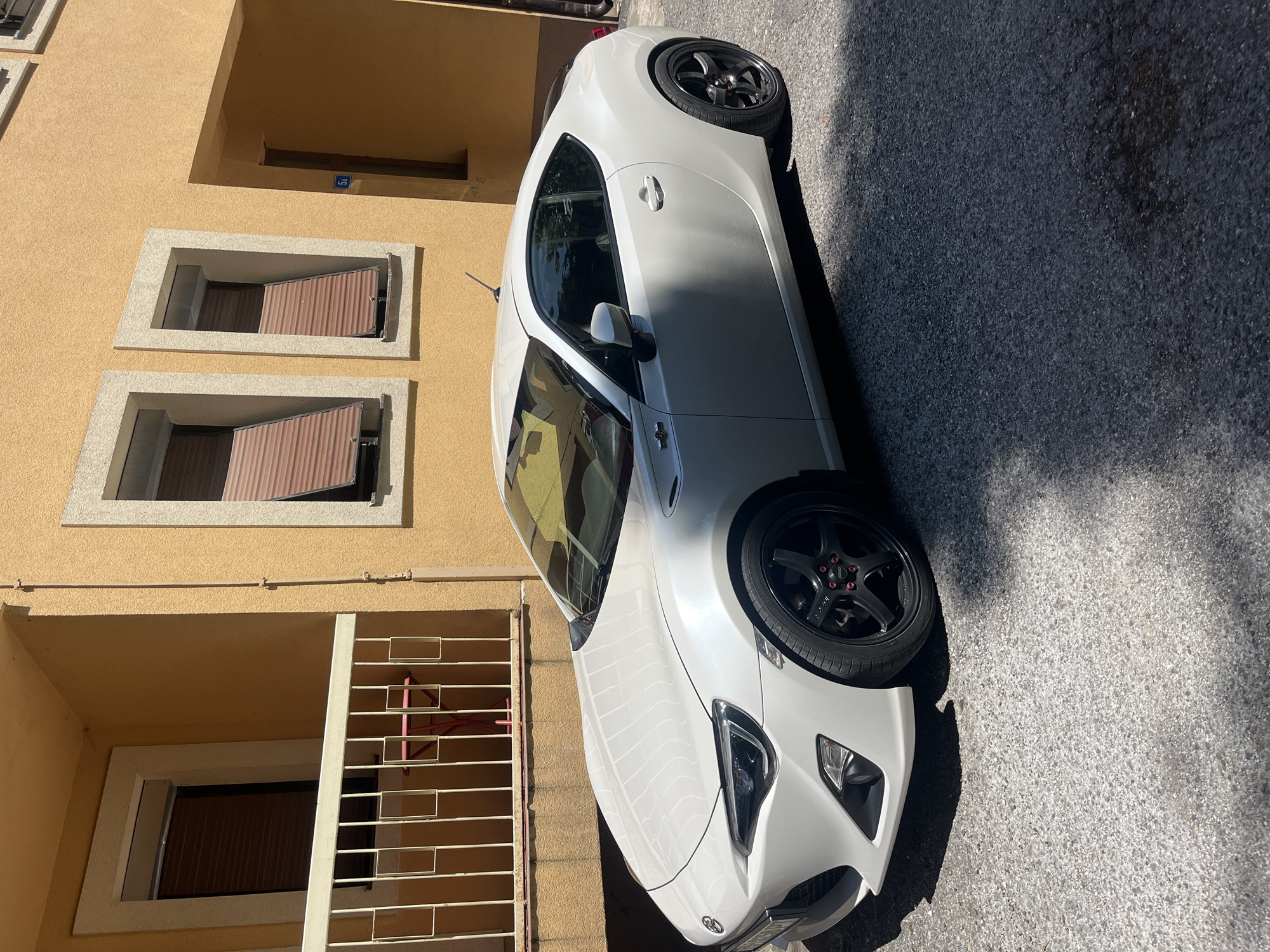 A white, two-door sports car is parked on an asphalt surface outside a building with yellow stucco walls. The car is positioned at an angle, facing towards the left of the frame. It features black alloy wheels with red lug nut caps. The building has several windows with brown shutters and a white railing on a balcony. The scene appears to be set in a residential area, likely a street or courtyard. The lighting suggests it is daytime with strong sunlight casting shadows. The location is specified as Vrela, Montenegro.