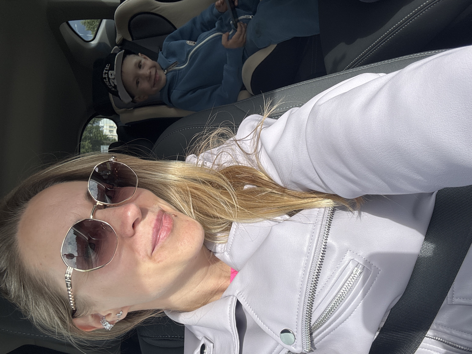 A selfie captures a woman and a young child inside a car, likely in Chaikovsky, Russia. The woman, in the foreground, has blonde hair, wears oversized sunglasses that reflect a building, and a light lavender leather jacket with prominent silver zippers and snap buttons, over a bright pink top. A dark seatbelt crosses her shoulder. Her expression is neutral with a slight smile. She has silver-toned earrings.
In the background, a young child, possibly a boy, is seated in a car seat. He wears a blue hooded jacket and a black and white baseball cap with "ATHLETIC" and possibly "28" or "EB" written on it. He is smiling broadly and looking towards the camera.
The car's interior features dark gray seats. Bright sunlight streams into the vehicle, creating strong highlights and shadows, indicating a sunny daytime. Through the car windows, blurred greenery and distant buildings are visible, suggesting an urban or suburban environment. The scene depicts a casual moment during travel, with both subjects seemingly relaxed and happy.