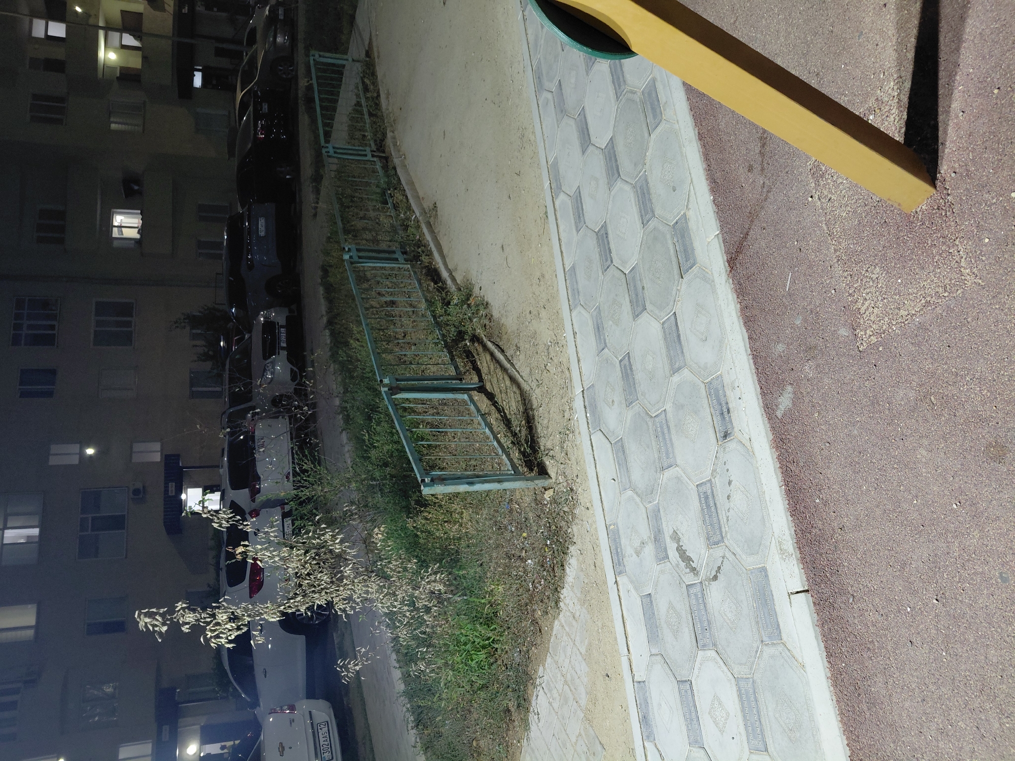 The image depicts an outdoor scene at night, likely in the city of Aktau, Kazakhstan. In the background, a multi-story apartment building is visible with illuminated windows. In front of the building, several cars are parked. A small tree with sparse, light-colored foliage stands to the left of the parked cars. A light blue metal fence runs horizontally across the mid-ground, separating the grassy area from a walkway. The walkway is paved with hexagonal gray tiles. In the foreground, a portion of a yellow playground structure is visible, casting a shadow on the reddish-brown ground cover. The ground cover appears to be a textured asphalt or similar material. The overall lighting suggests artificial illumination from streetlights or building lights.