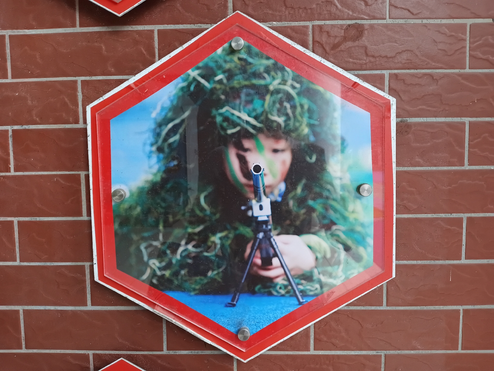 A photograph of a child in camouflage holding a gun on a tripod, displayed in a red and white hexagonal frame mounted on a brick wall. The location is Shunde District, China.