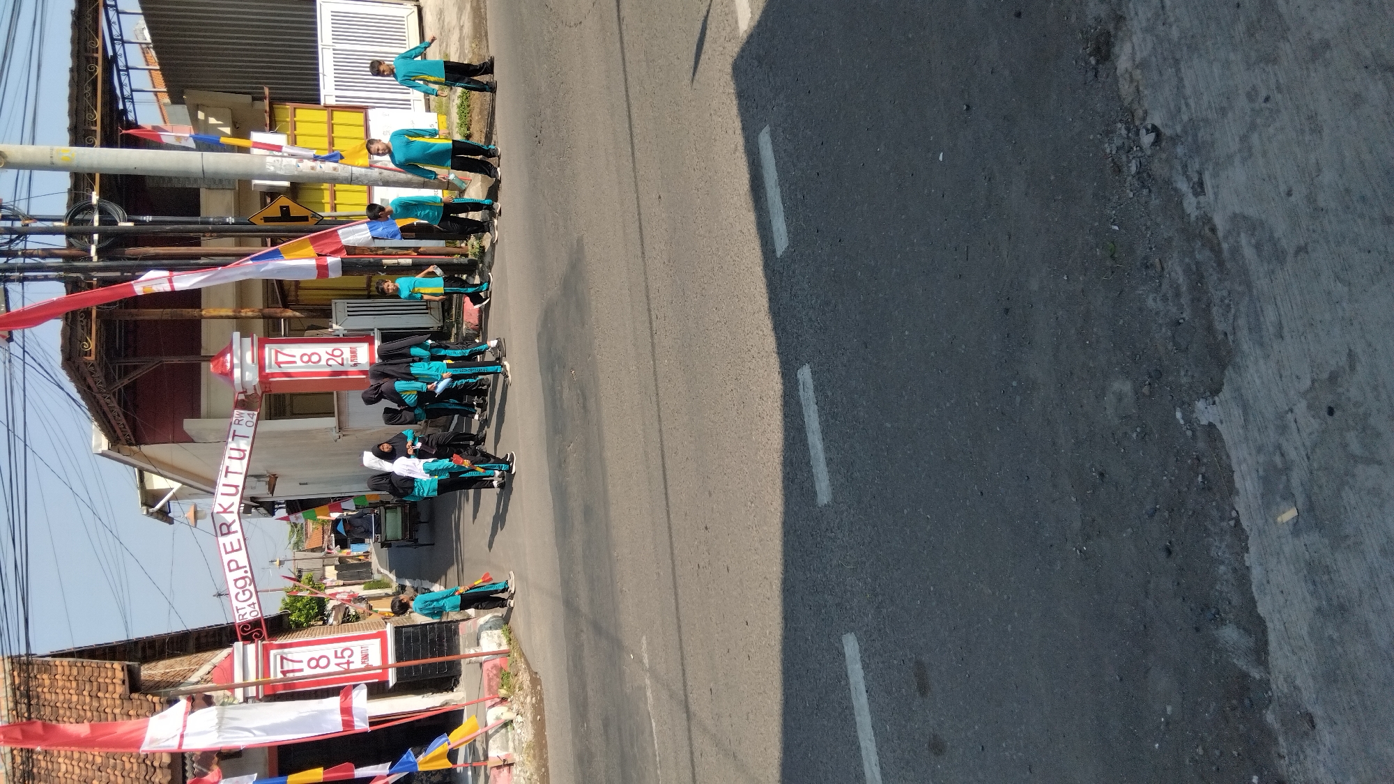 In the city of Proyonanggan Tengah, Indonesia, a street scene unfolds under a clear blue sky. A group of children, dressed in teal and black athletic uniforms, stand on the asphalt road. Some hold small red and yellow flags. To the left, a white archway stands, adorned with red and white flags and signs displaying numbers and text. A horse-drawn cart is visible further down the street. Buildings line the street, some with red-tiled roofs, and a tall utility pole with wires stands to the right. The road has white lane markings.