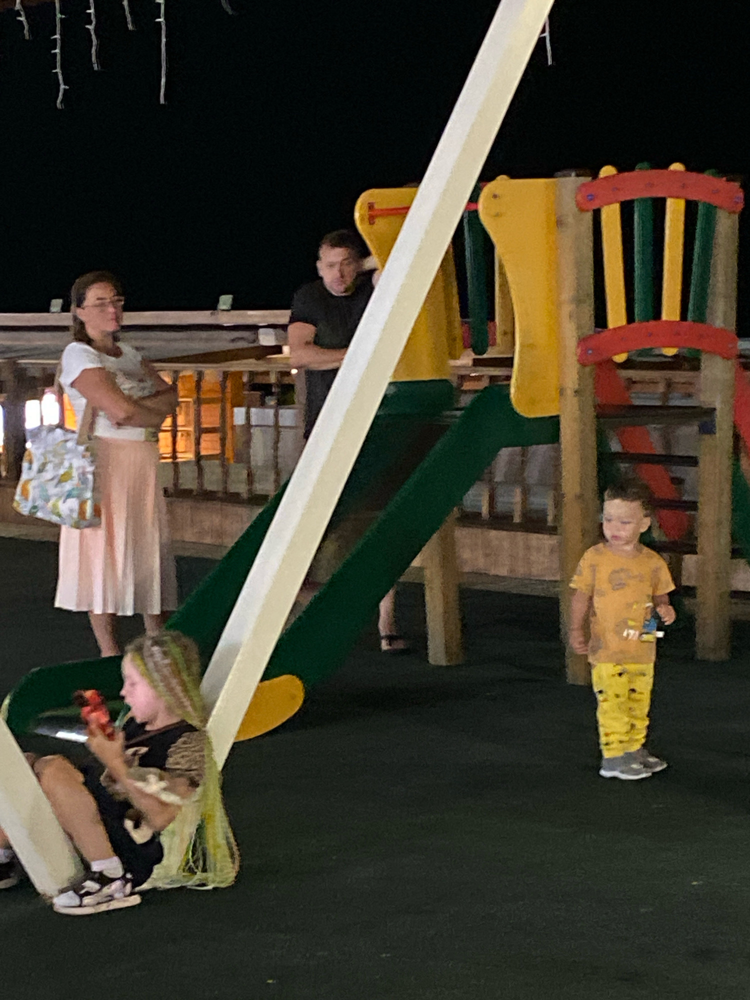The image depicts an outdoor playground scene at night. In the foreground, a young girl with blonde and green-streaked hair is sitting on the ground, looking at a red object in her hands. She is wearing a black and gold shirt and black shorts with white sneakers. Behind her, a young boy in a yellow t-shirt and yellow pants with cartoon characters is standing.
In the background, two adults are visible. A woman in a white t-shirt and a long, light pink skirt is standing with her arms crossed. She is carrying a patterned bag over her shoulder. To her right, a man in a black t-shirt is standing with his arms crossed.
The playground structure is prominent, featuring yellow, green, and red elements, including a slide and ladder. The ground surface is dark, likely rubberized safety surfacing. String lights are visible hanging in the dark sky above the playground. The overall setting suggests a leisure area in a public space. The context of Sochi, Russia, is provided, indicating the geographical location of the scene.