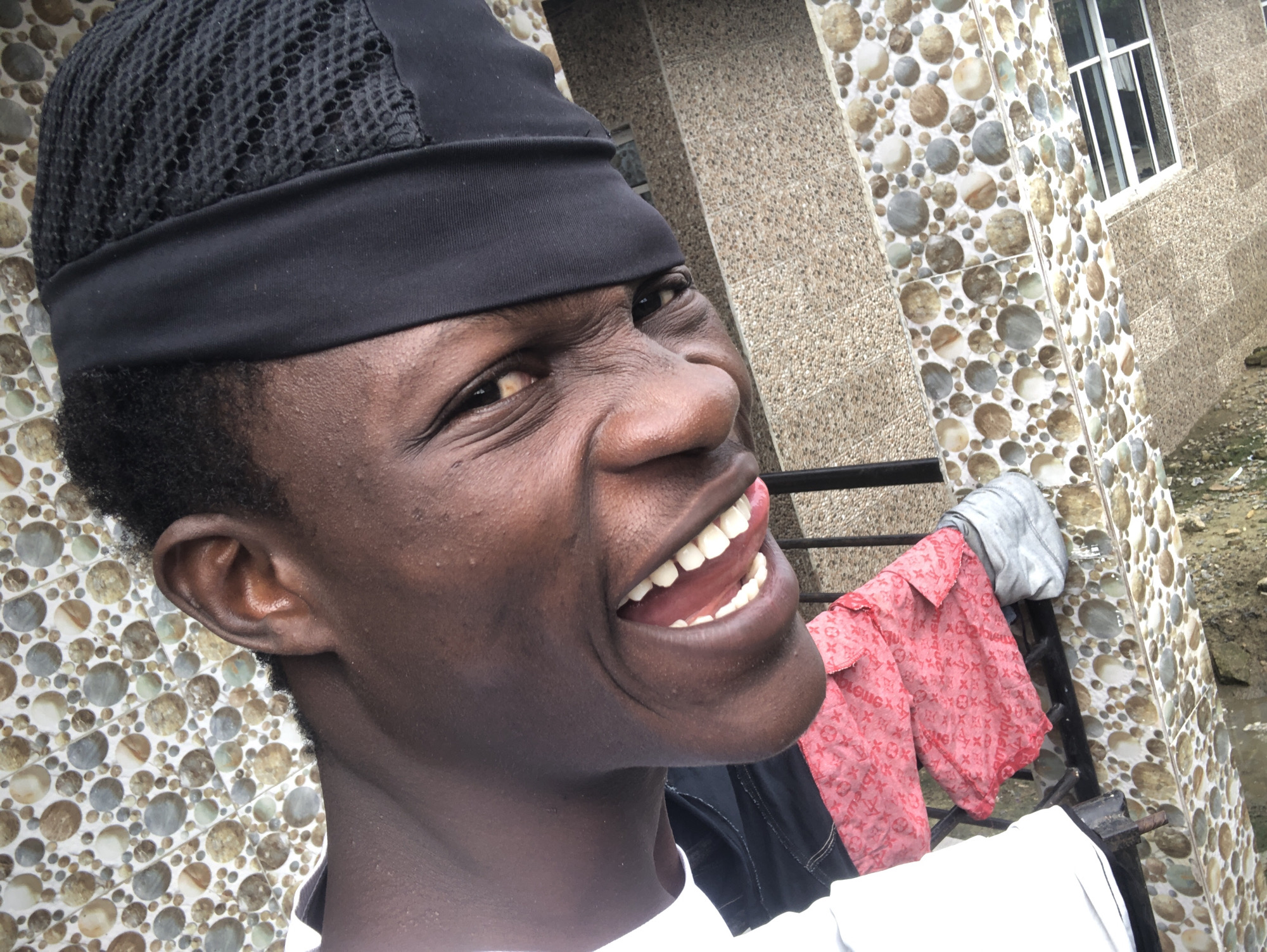A medium-shot, low-angle, selfie-style photograph captures a Black male wearing a black skullcap. He is looking directly at the camera with a wide, toothy grin, revealing his tongue and uvula. His eyes are squinted, and his left eyebrow is raised, suggesting amusement. The background is a textured wall with circular mosaic tiles in various shades of brown and beige, with a vertical strip of lighter-colored, rectangular tiles. To the right of the man, a metal railing is visible, with a folded gray garment and a red garment with a repeating pattern of "LV" logos hanging from it. The scene appears to be outdoors, possibly in Ado, Nigeria, given the style of architecture and the presence of drying clothes, a common sight in residential areas. The lighting is bright, casting shadows on the man's face and highlighting the texture of his skin.
