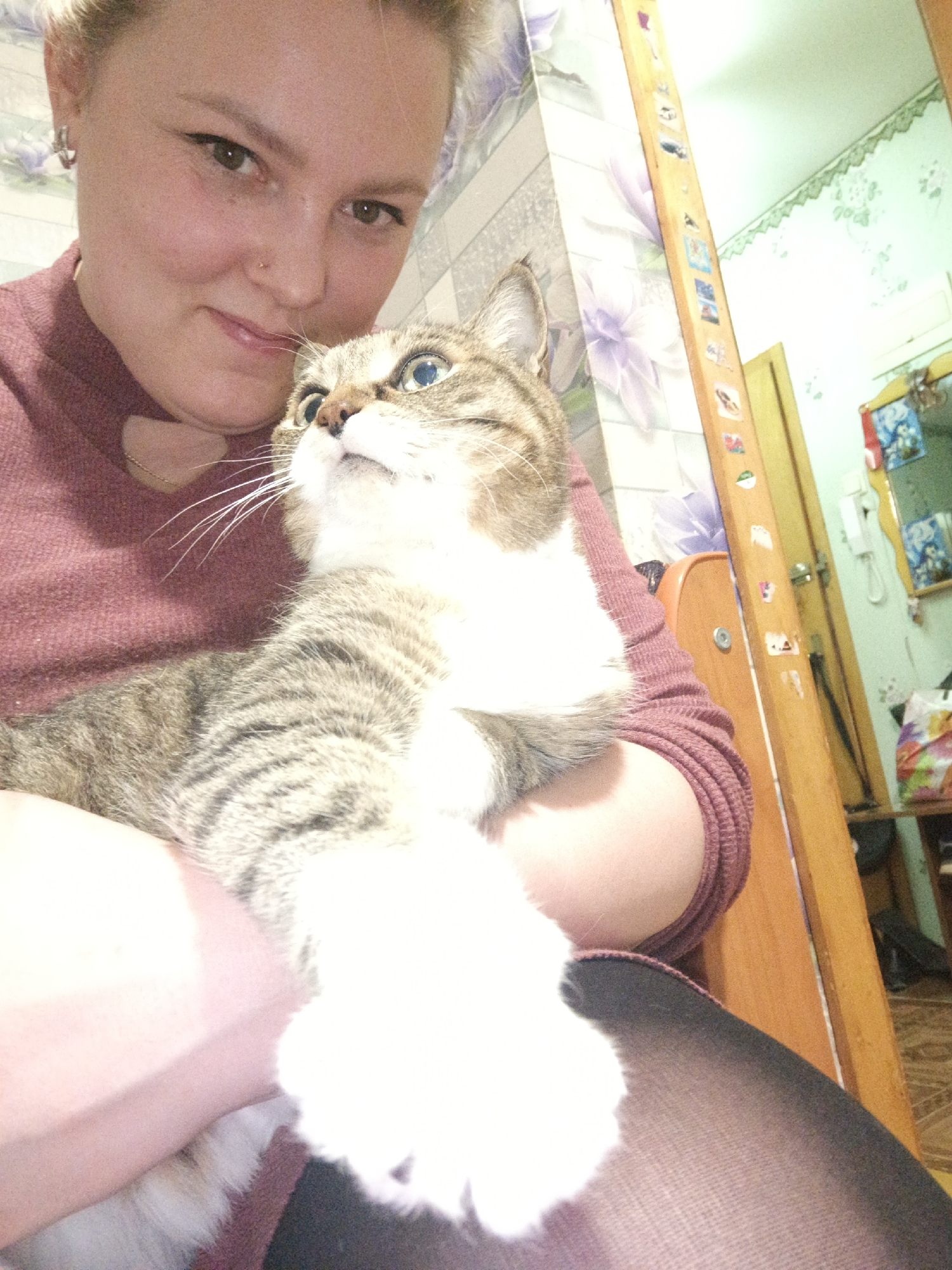 This media file captures a close-up, casual portrait of a woman holding a cat in an indoor setting in Марковский, Russia.
The central subjects are a young woman and a domestic cat. The woman, who appears to be in her 20s or 30s, has light-colored hair pulled back and is smiling gently at the camera. She has brown eyes, a small nose piercing, and an earring. She is wearing a maroon or deep red knitted sweater with a subtle cutout detail near the collarbone and dark patterned leggings.
The cat, nestled comfortably in her arms, is a tabby with distinctive brown, grey, and black stripes, and large white patches on its chest and front paws. It has striking light blue or greenish-blue eyes and is looking intently upwards and slightly to the right, away from the camera. Its whiskers are prominent.
The activity depicted is a warm, affectionate moment between a pet owner and her cat, suggesting a bond of companionship. The woman is cradling the cat, which seems relaxed and curious.
The setting is an indoor room, likely a home, with varied wallpaper patterns visible in the background. One wall features a light-colored paper with soft purple floral designs, while another has a lighter green or blue pattern. To the right, a tall wooden frame, possibly part of a mirror or door, is adorned with numerous small, colorful stickers or magnets; the specific content of these is not legible. Further in the background, a light-colored wooden door and a table holding various household items, including bags and another small mirror, can be seen. The lighting suggests an indoor daytime scene or a well-lit room. The overall mood is personal and heartwarming. No legible text is visible in the image.