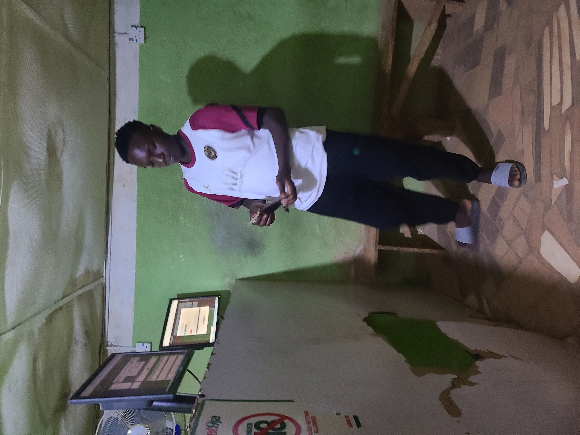 A young man stands in an internet cafe in Unknown, Unknown, focused on something he holds in his hands. He wears a white and burgundy t-shirt, black pants, and gray slides. He is positioned between two computer monitors and a counter with advertising. The cafe has green walls. The floor is tiled with a brown and white pattern.