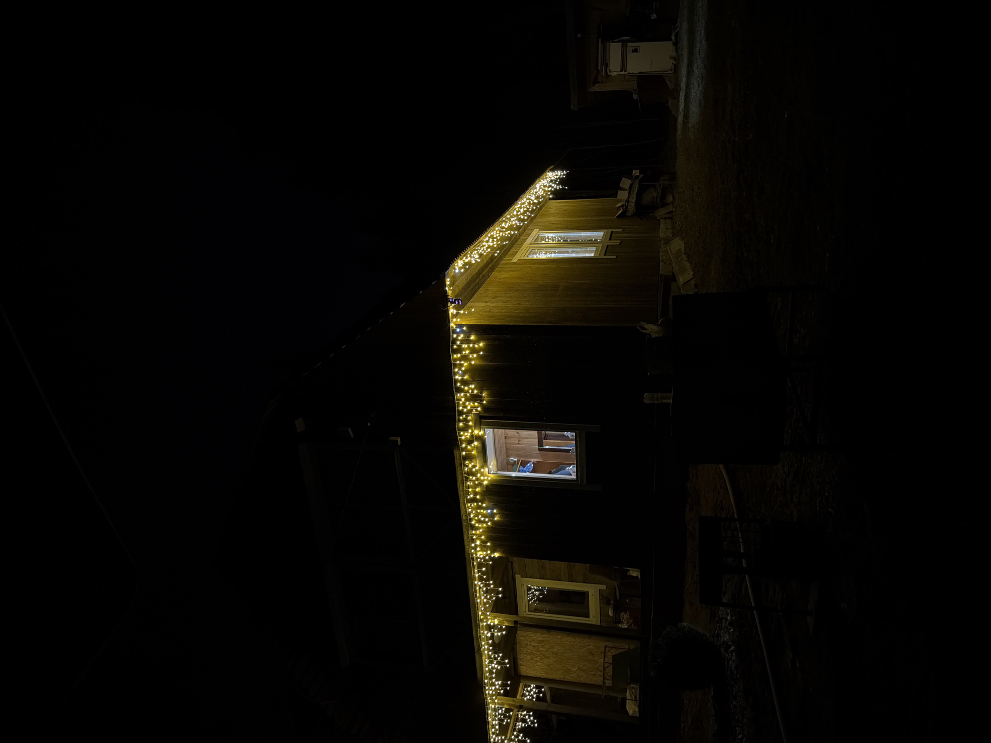 A low-angle shot of a building at night, decorated with string lights. The building's exterior is wooden, with windows illuminated from the interior. The background is dark, with minimal visibility of the surroundings. The location is inferred to be Starosel'e, Russia. No people are visible.
