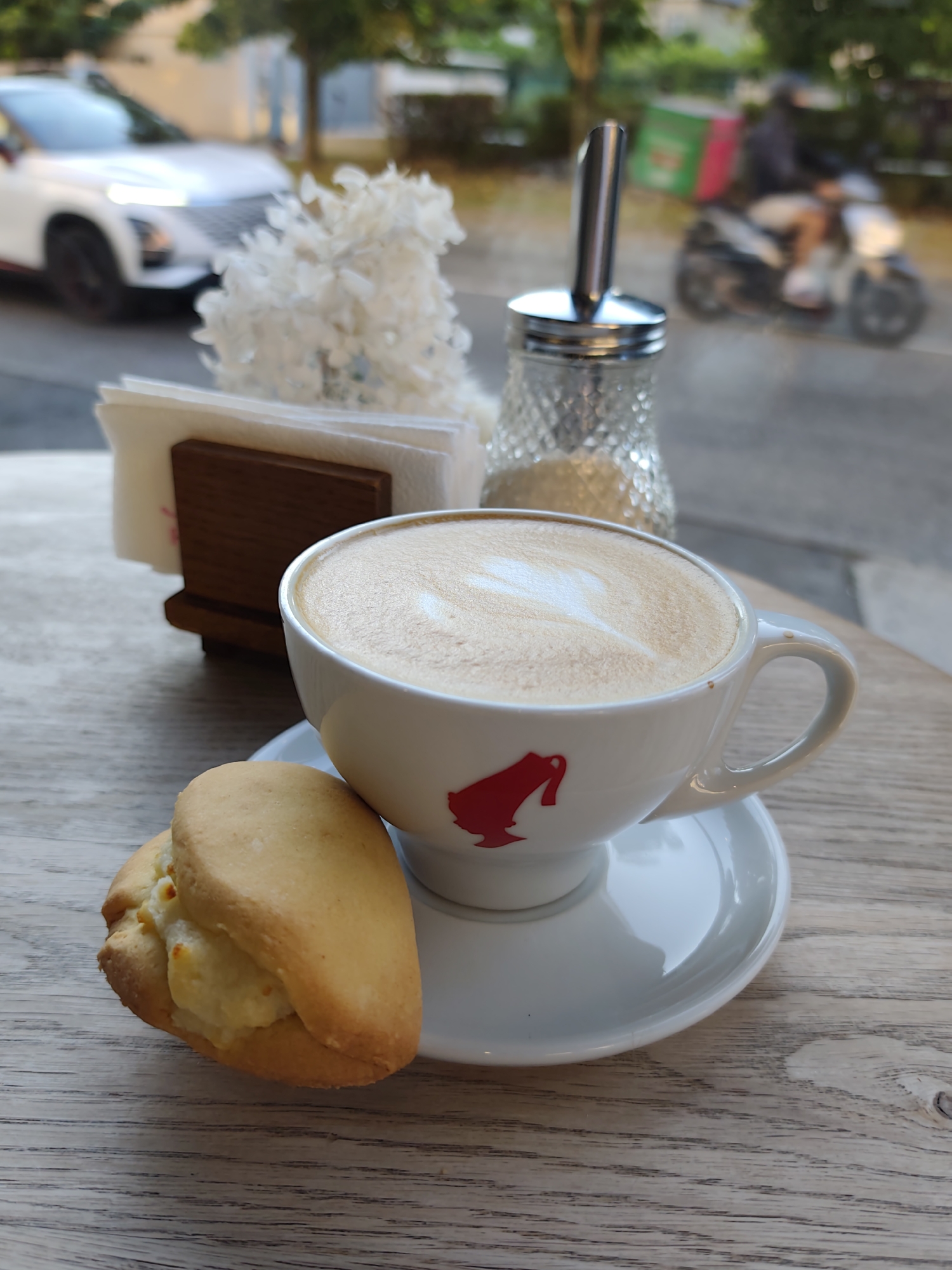 A close-up, eye-level shot captures a coffee cup with latte art and a cookie on a wooden table. The coffee is in a white cup with a red emblem, sitting on a saucer. Beside the cup is a golden-brown cookie with a creamy filling. In the background, a sugar dispenser is visible, along with a wooden napkin holder filled with white napkins and a decorative white floral arrangement. Through a window or glass partition, an out-of-focus urban scene unfolds. A white car is parked on the left, and a person on a scooter with a delivery box is in motion on the right. The environment suggests an outdoor seating area of a cafe or restaurant. The metadata indicates the image was taken in Sochi, Russia, on May 20, 2024.