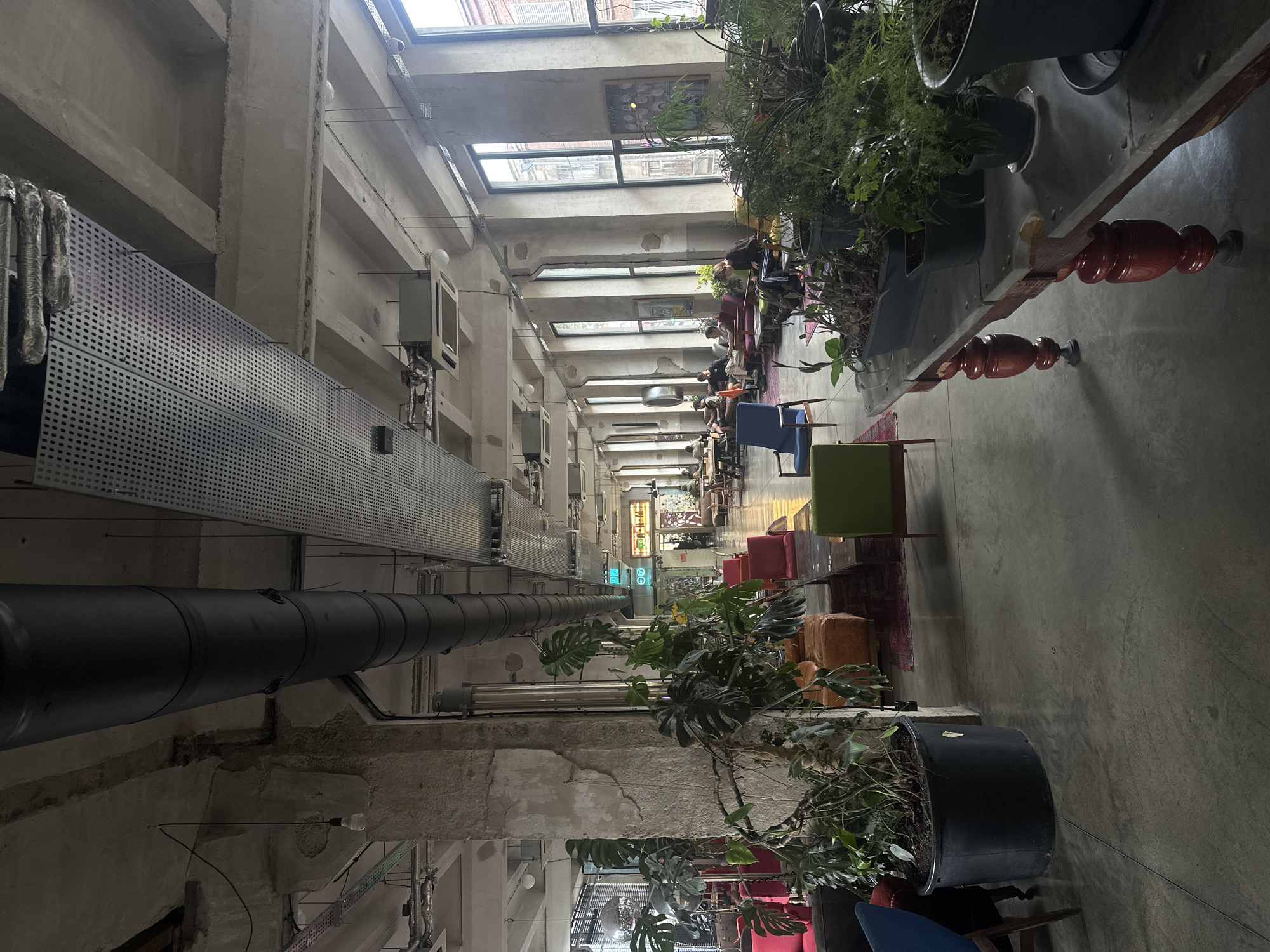 This image shows the interior of a cafe or co-working space in Tbilisi, Georgia, characterized by its industrial-chic aesthetic. The space features exposed concrete ceilings, walls, and floors, with large, tall windows allowing natural light to flood the area. Prominent industrial elements include large black pipes running along the ceiling and metal conduits and ventilation systems.
The furniture is a mix of modern and eclectic styles. Various seating areas are arranged throughout, including plush red armchairs, orange poufs, and blue and green accent chairs. Several large potted plants, most notably a substantial monstera, are strategically placed, adding a touch of greenery to the urban environment. A long, rustic table with turned wooden legs, holding several potted plants, is visible in the foreground.
In the background, patrons are seated at tables, engaged in activities such as working on laptops or conversing. The overall atmosphere appears relaxed and communal. The lighting is a combination of natural light from the windows and overhead artificial lighting, including bare bulbs and modern fixtures. The architectural style and the presence of plants suggest this is a contemporary establishment within the city, likely a popular spot for freelancers and social gatherings.