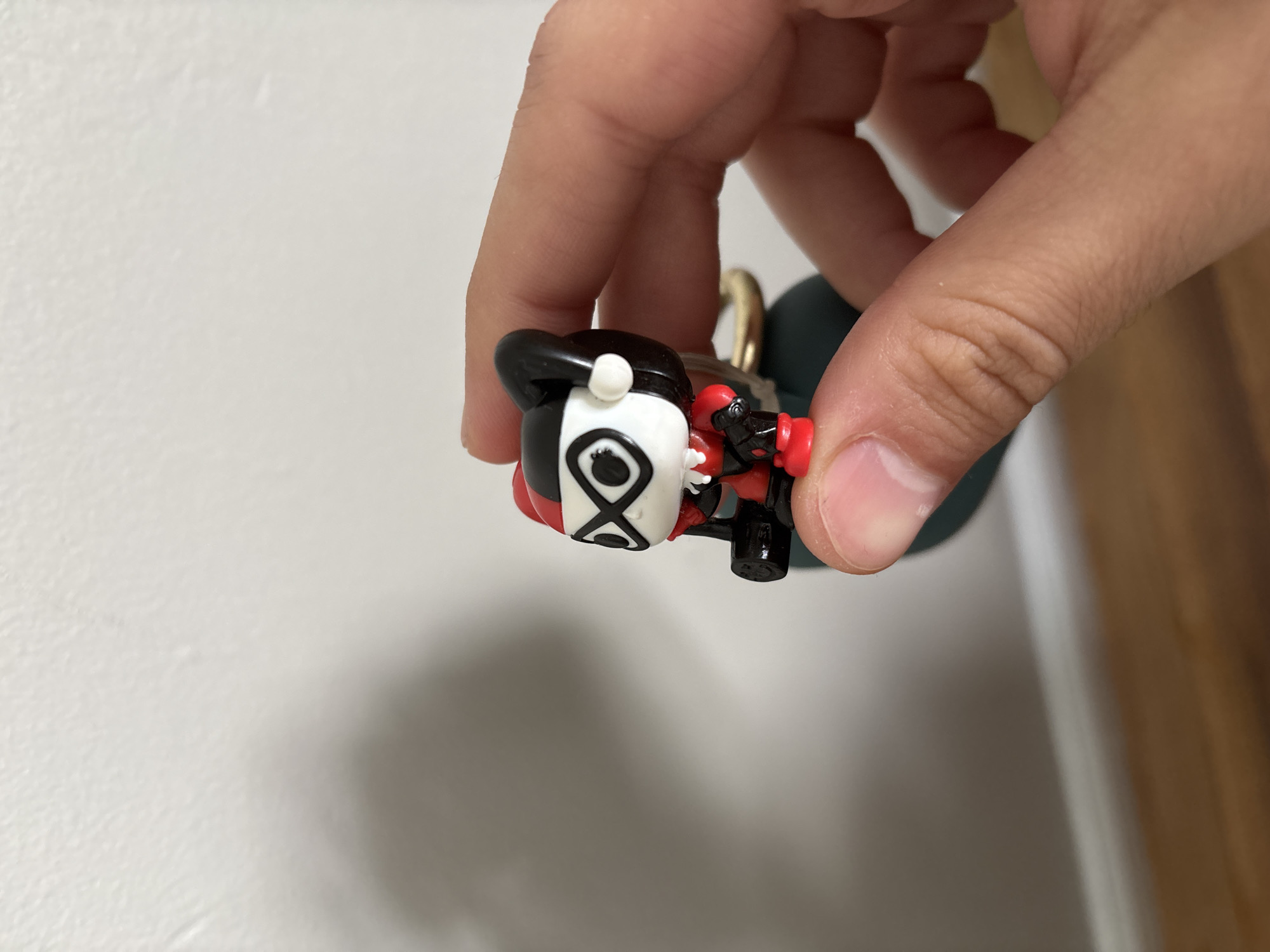 A person is holding a small Harley Quinn figurine in their fingers. The figurine is primarily black, white, and red, with details suggesting the character's jester-like appearance. The background consists of a white wall and a wooden surface, likely a door or trim. The location is Franklin Township, United States of America, based on image context clues.