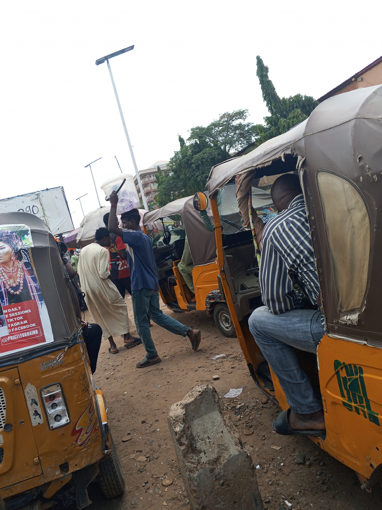The image shows a dusty scene in Abuja, Nigeria, featuring a row of yellow "keke" or auto-rickshaws. The ground is unpaved. Several people are visible; one man carries a clear plastic bucket on his head, while another sits inside one of the auto-rickshaws. Advertising posters are affixed to some of the vehicles. A street lamp is visible in the background. The buildings in the background are multi-story.