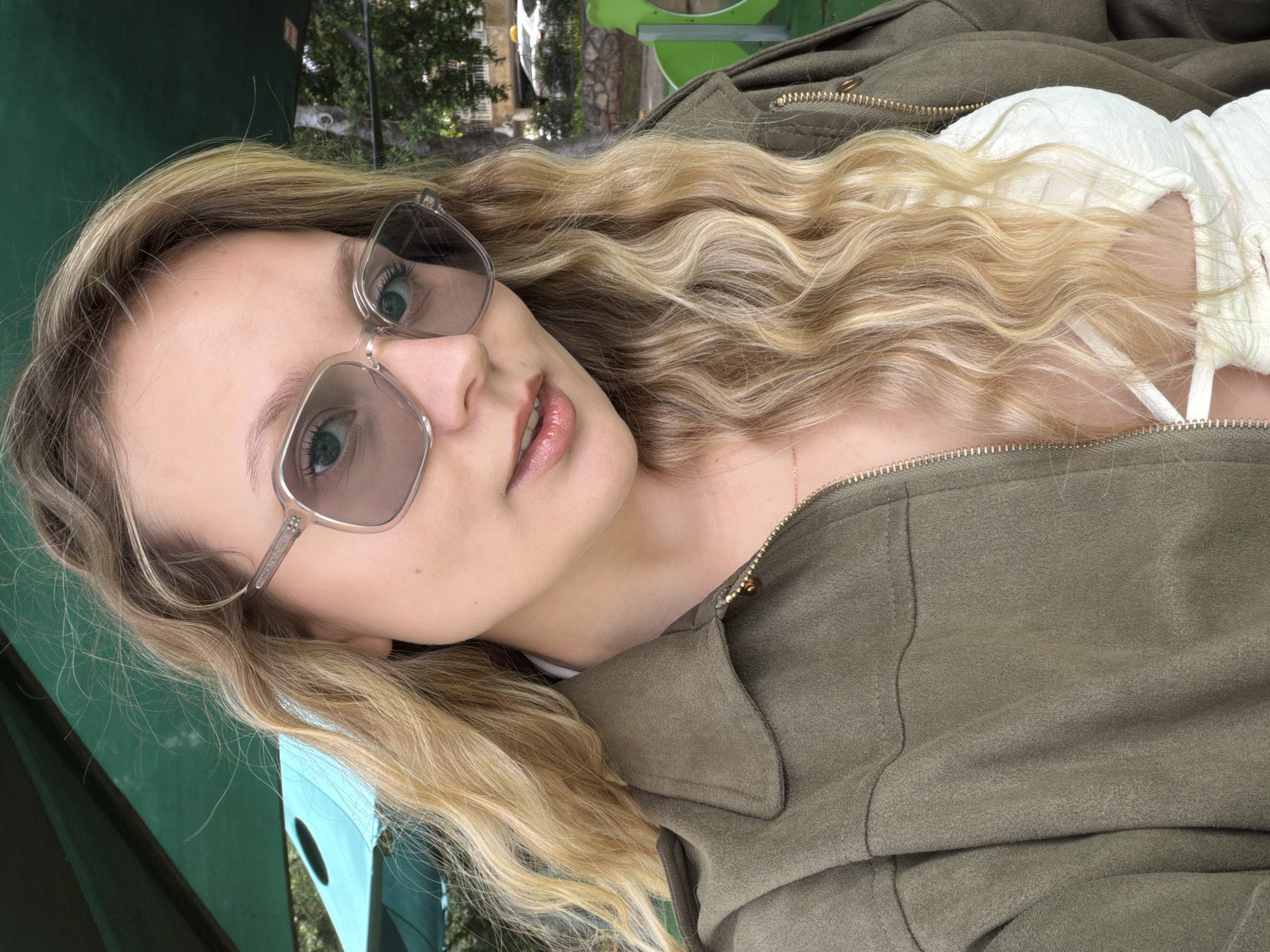 A fair-skinned female subject with long, wavy blonde hair is depicted in a medium shot, looking directly at the viewer. She is wearing clear-framed, gray-tinted sunglasses, revealing her green eyes. Her lips are slightly parted, exhibiting a light, glossy finish. She wears an open, olive-green, suede-like jacket with a gold-colored zipper partially visible on the right. Beneath the jacket, a white, textured garment, possibly a top or bra, is visible, featuring a cut-out design on the chest area. A delicate gold-colored chain necklace with a small pendant rests on her skin.
The scene is outdoors during daylight, consistent with a public park or playground environment in Tel-Aviv. In the immediate background, a dark green canopy is overhead, and to the left, a portion of a lighter blue canopy is visible. To the right, green playground equipment with yellow accents is partially visible. Further in the background, out of focus, are green trees, a light-colored vehicle (possibly a white taxi with a yellow roof sign), and a stone wall or barrier, indicative of an urban setting. No other individuals or specific actions beyond the subject posing are discernible.