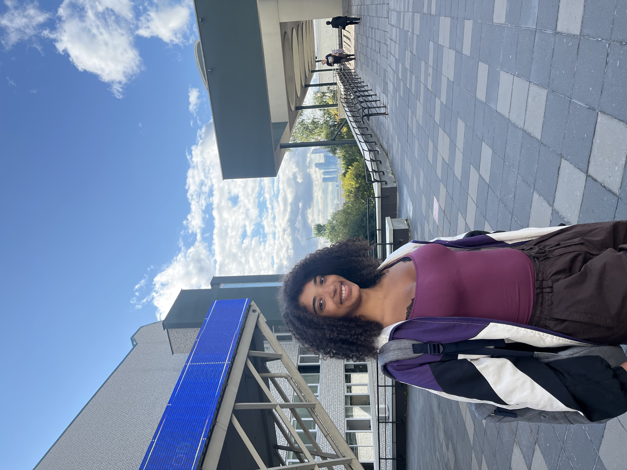 A young woman with curly dark hair, wearing a purple tank top, dark brown pants, and a purple, white, and black jacket, smiles at the camera while standing on a paved walkway. She carries a gray backpack. Behind her, a building with a light gray facade and blue accents is visible. The walkway extends into the distance, lined with metal railings and leading towards a covered area. In the background, trees and a distant city skyline with skyscrapers can be seen under a bright blue sky with scattered clouds. The scene appears to be an outdoor public space within a city. The architectural style of the buildings and the presence of numerous skyscrapers in the background are consistent with a location in Moscow, Russia.