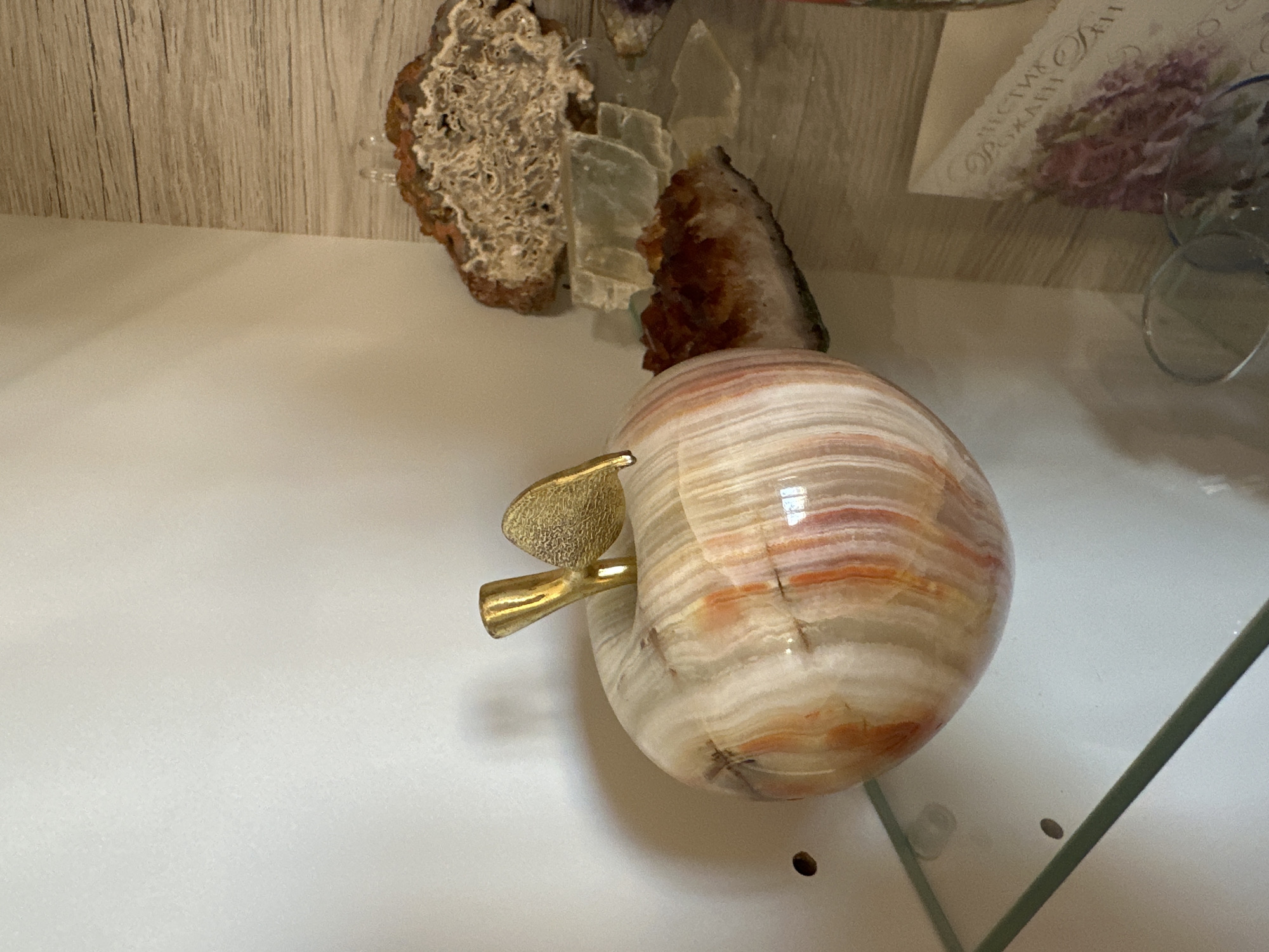 The image shows a close-up of an object resembling a striped stone apple with a gold-colored stem and leaf. The apple is placed on a white surface, likely a shelf. In the background are other geological specimens or decorative objects, including what appears to be an agate slice and possibly a birthday card in Bulgarian. The scene appears to be indoors, potentially within a collector's display cabinet or on a shelf in a home in Sofia, Bulgaria.
