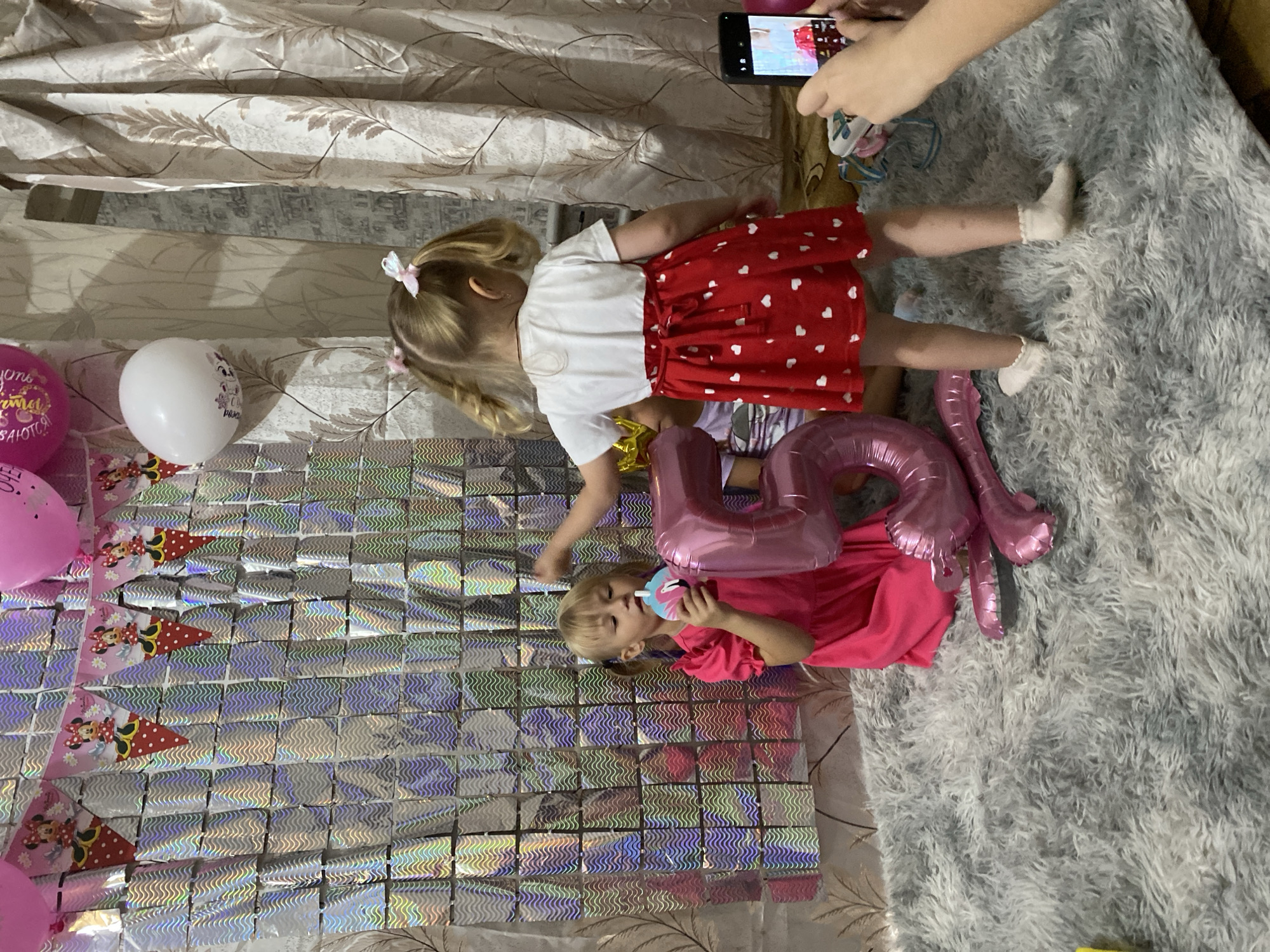 In a room decorated for a child's birthday, two young girls are the focus of the scene. The setting appears to be indoors, possibly a living room, with a fluffy grey rug and a wall adorned with shimmering, reflective squares. A patterned curtain hangs in the background.
The older girl, on the left, is wearing a white t-shirt and a red skirt with white hearts. Her hair is pulled into two pigtails, each adorned with a small white bow. She is facing away from the camera, her back towards the viewer.
The younger girl, on the right, is wearing a bright pink dress and is sitting on the rug. She is holding a pink and blue party blower to her mouth and appears to be blowing into it. She has blonde hair and is looking towards the camera.
Between the two girls are large pink foil balloons shaped like the numbers "5" and "0", suggesting a 50th birthday, though the presence of young children might indicate a party for someone else with these numbers as a theme, or possibly a 5th birthday of one of the children. There are also pink and white balloons scattered around, including one with a Minnie Mouse design and another plain white one. Party banners featuring Minnie Mouse are also visible on the decorated wall.
A hand holding a smartphone is visible in the upper right corner, capturing the moment, indicating that a photo or video is being taken. The overall atmosphere seems festive and joyful, suggesting a birthday celebration. The image is taken in Маринопіль, Ukraine.