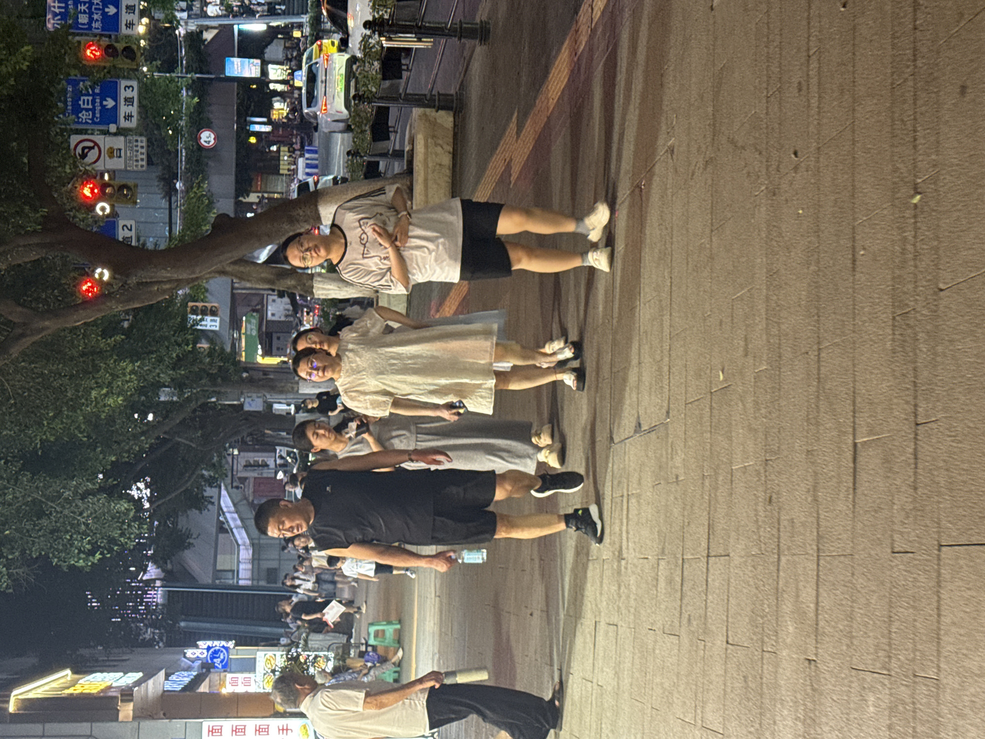 A group of people are walking on a tiled sidewalk in Yuzhong District, China. The scene appears to be at night or in the evening, with artificial lighting illuminating the area. The people are dressed in casual attire, suggesting they are likely enjoying a walk or outing.
In the foreground, a man in a black sleeveless shirt and black shorts is walking towards the viewer, holding a bottle of water in his right hand. To his left, an elderly man in a light-colored short-sleeved shirt and dark pants is walking away from the viewer. Behind the man in the black shirt, a woman in a grey dress and a woman in a light-colored dress are walking together. Further back, another woman is visible.
The sidewalk is paved with rectangular tiles. Behind the sidewalk, there are trees, buildings with illuminated signs, traffic lights, and cars on the road. The presence of streetlights and illuminated building signs indicates an urban setting. The overall atmosphere is that of a busy street scene at night.