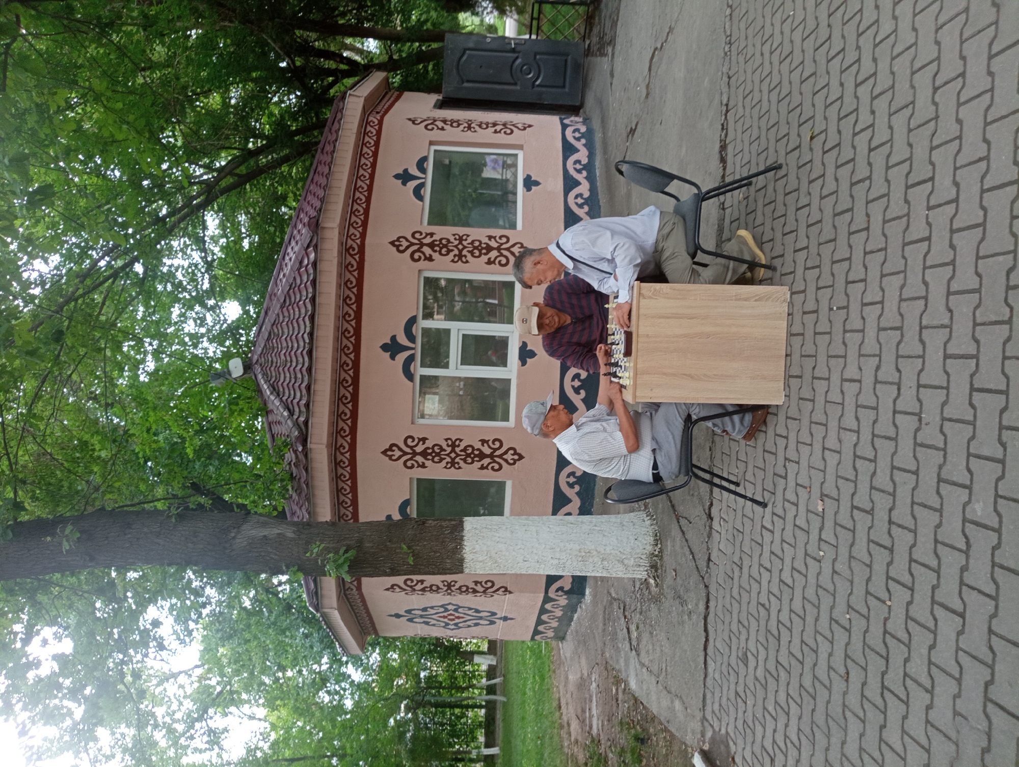 In Shymkent, Kazakhstan, three men are playing chess at a small wooden table outdoors. They are seated on black chairs in front of a circular building with a reddish-brown tiled roof and pink walls adorned with decorative patterns. The building has windows and a dark door on the right side. The men are engaged in the game, with chess pieces visible on the board. The scene is set amidst trees with green foliage and a paved ground made of interlocking grey bricks. The overall atmosphere suggests a leisurely activity in a park-like setting.