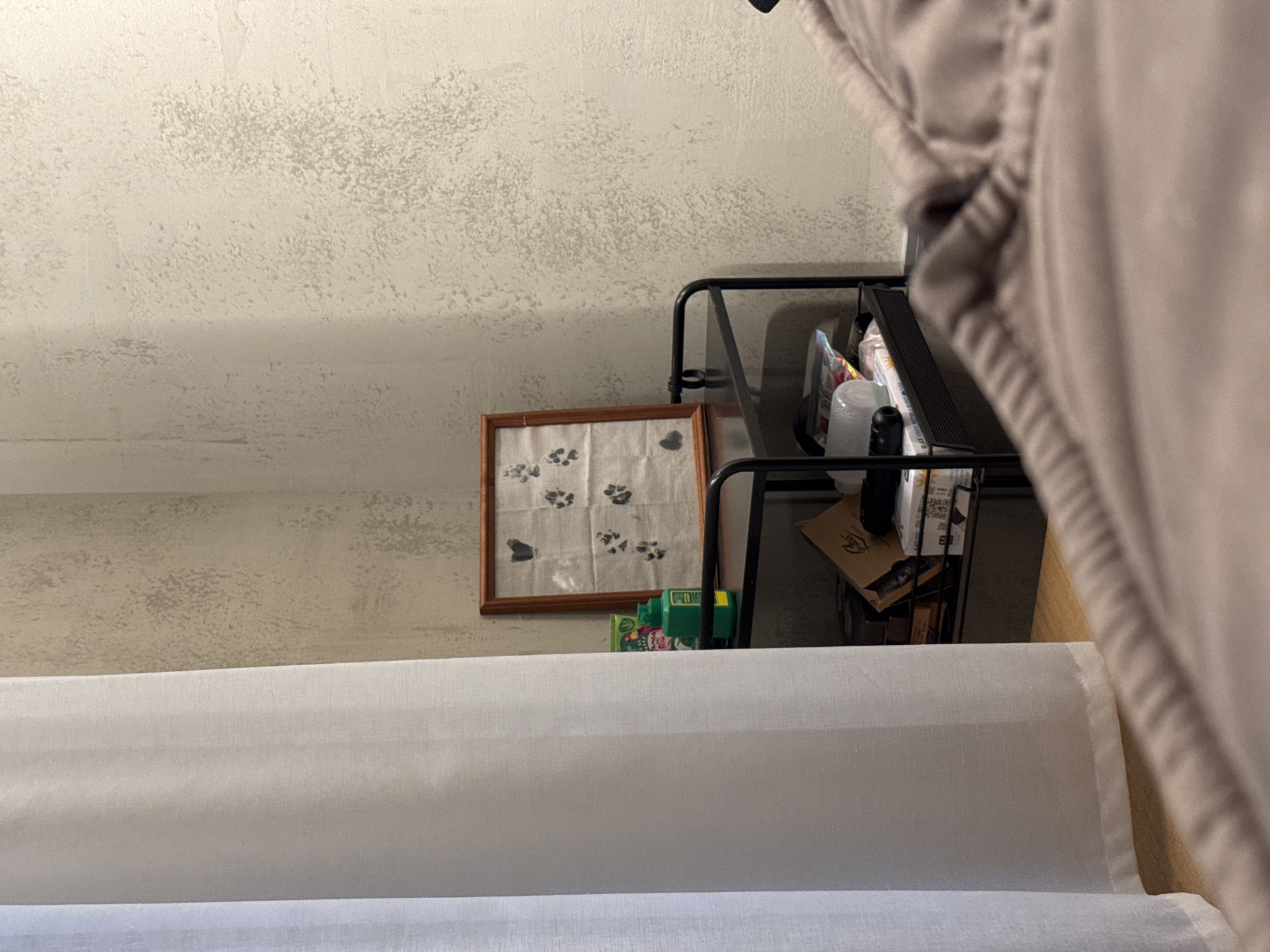 The image depicts an indoor scene with a focus on a shelf unit and wall textures. On the top shelf of a black metal shelf unit, a framed piece of art is visible, featuring a pattern of paw prints. To the left of the artwork, a green bottle with a white cap is present. The shelf unit holds various items on its lower shelf, including what appears to be stacked boxes and a clear plastic container. The background consists of textured walls, one with a light, rough finish and another appearing to be a corner. A white curtain hangs on the left side of the frame, partially obscuring the shelf unit. The foreground shows a blurred section of what seems to be bedding or upholstery. The scene does not provide explicit clues to its exact location within Бараўлянскі сельскі Савет, Belarus.