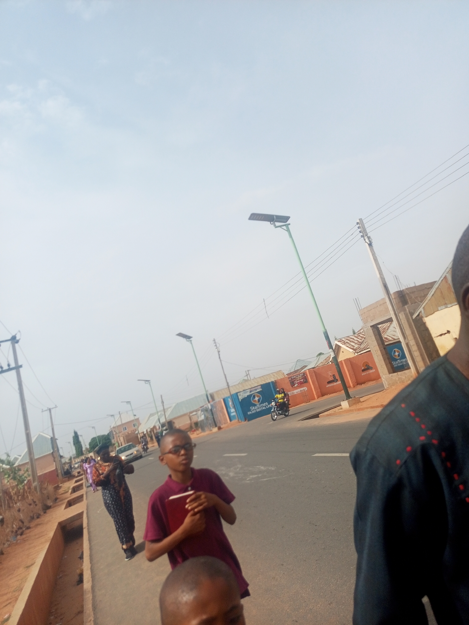 The image depicts a street scene in Tempure, Nigeria. The street is paved and lined with sidewalks and buildings. Several people are walking on the sidewalk, including a boy holding a book, a woman in a patterned dress, and a man whose partial figure is visible on the right. A motorcycle with two passengers is driving on the street. There are several utility poles and solar-powered street lights along the road. Some buildings have signs for "Startimes." The sky is overcast.