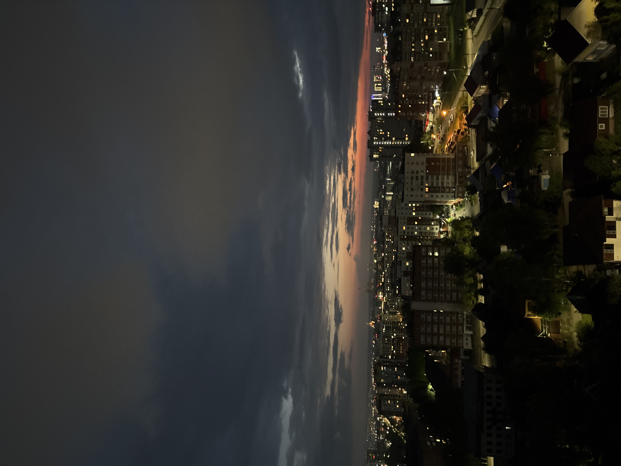 This image captures a twilight view of a city, likely Izhevsk, Russia. The sky is dominated by dark, heavy clouds with a sliver of orange and pink sunset visible on the horizon, hinting at the end of daylight. Below the sky, a sprawling urban landscape unfolds, characterized by densely packed apartment buildings illuminated by numerous lights. The buildings vary in height and architectural style, with many featuring brick facades and numerous visible windows. In the foreground and middle ground, there are also lower-rise buildings and what appear to be trees, creating a contrast between the high-rise apartments and the more suburban or older parts of the city. Streets are visible, with some car lights suggesting ongoing activity. The overall atmosphere is nocturnal and somewhat moody, typical of a city transitioning from day to night under overcast skies. No people are discernible in the image.