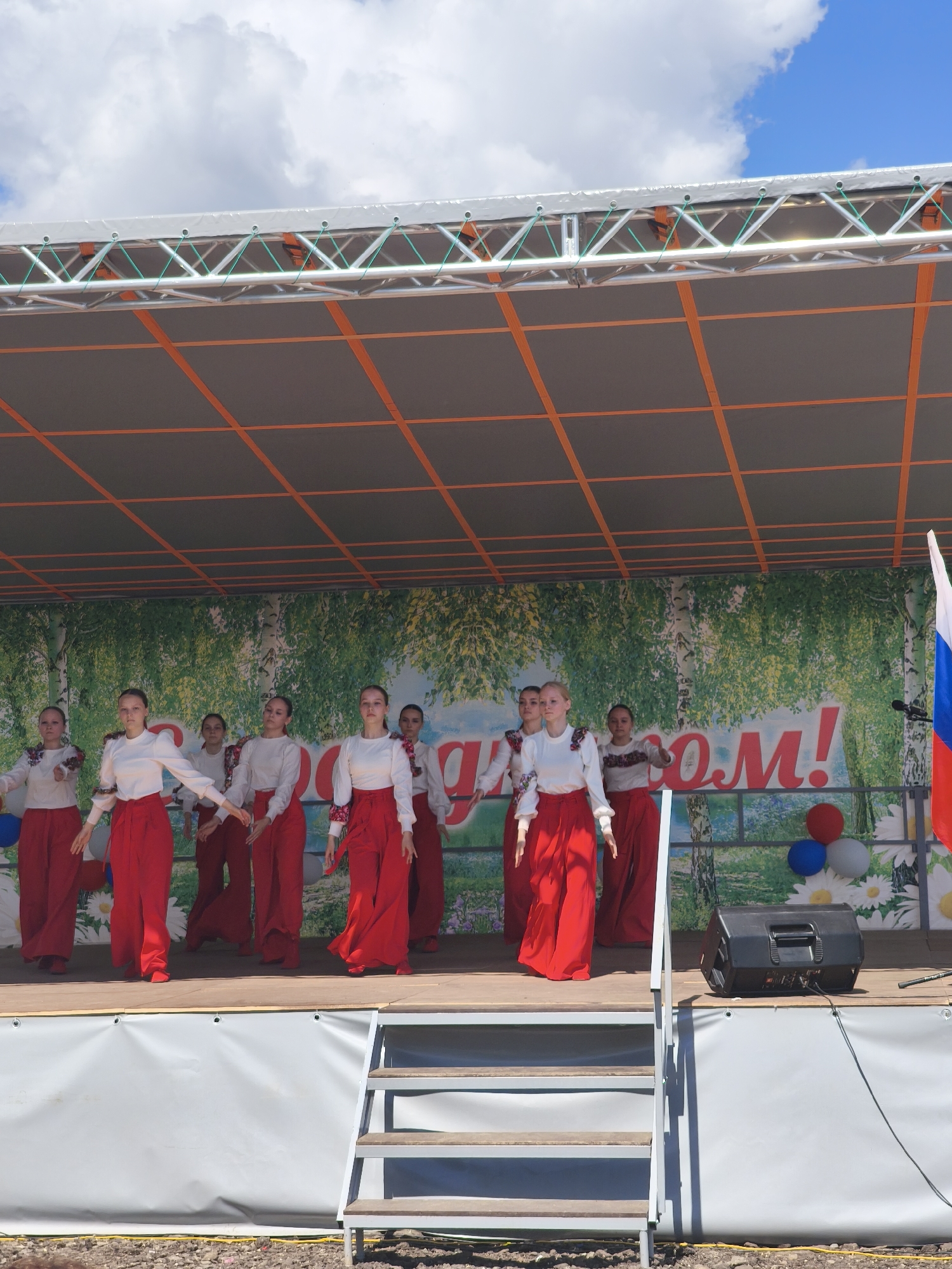 The image shows a stage performance in Kady, Russia. A group of young women in traditional red and white attire are dancing on a stage set up outdoors. The backdrop features a print of birch trees and a banner with large red lettering that appears to read "ПОБЕДОМ!". The stage is covered by a canopy supported by metal trusses, and the sky above is blue with white clouds. To the right of the stage, a Russian flag on a pole is visible, along with some balloons and a speaker. A set of metal stairs leads up to the stage on the right side of the image. The ground in front of the stage appears to be covered with gravel or dirt.