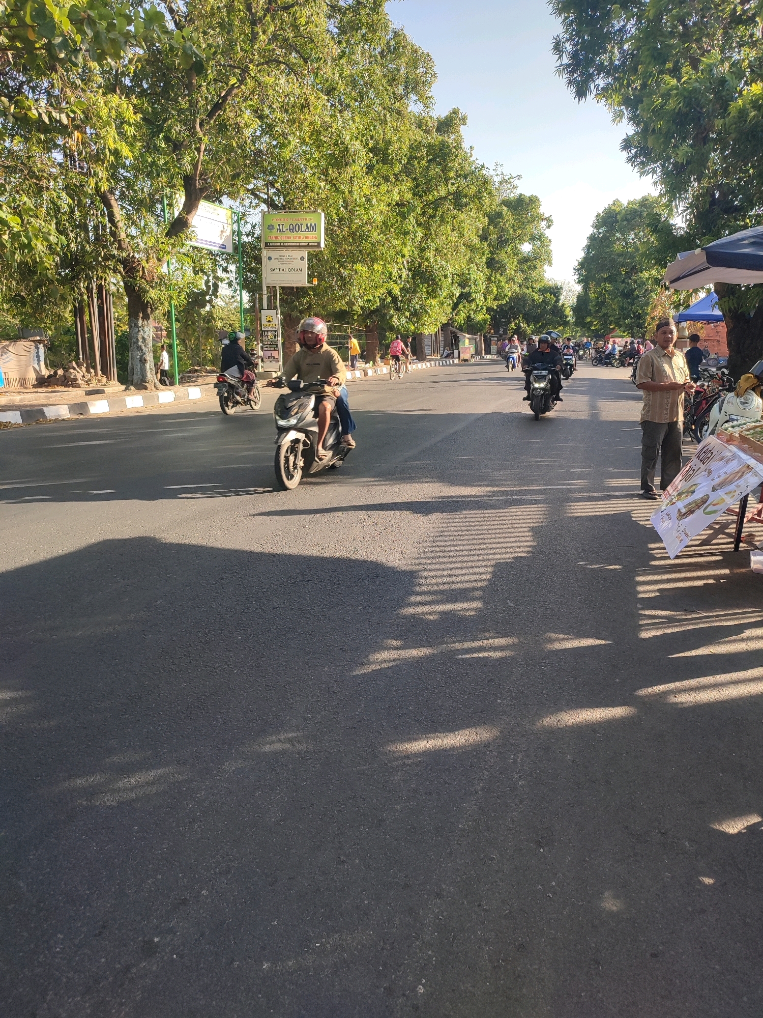 This is a full-frame, eye-level, outdoor, vertical, daytime photograph of a street scene in the city of Watubelah, Indonesia. The image captures a road with several vehicles and pedestrians. On the left side of the road, there is a sidewalk with a few people walking and a sign that reads "AL-QOLAM." The road itself is paved and has white lines marking lanes. A person on a scooter is in the foreground, heading away from the camera. Behind them, other motorcycles and a few bicycles are visible. On the right side of the road, a man in a light-colored shirt and dark pants stands next to a small stall with a sign that appears to advertise food. The background is filled with lush green trees lining the street, and the sky is clear and bright. The overall impression is of a busy, sunny day in a tropical urban environment.