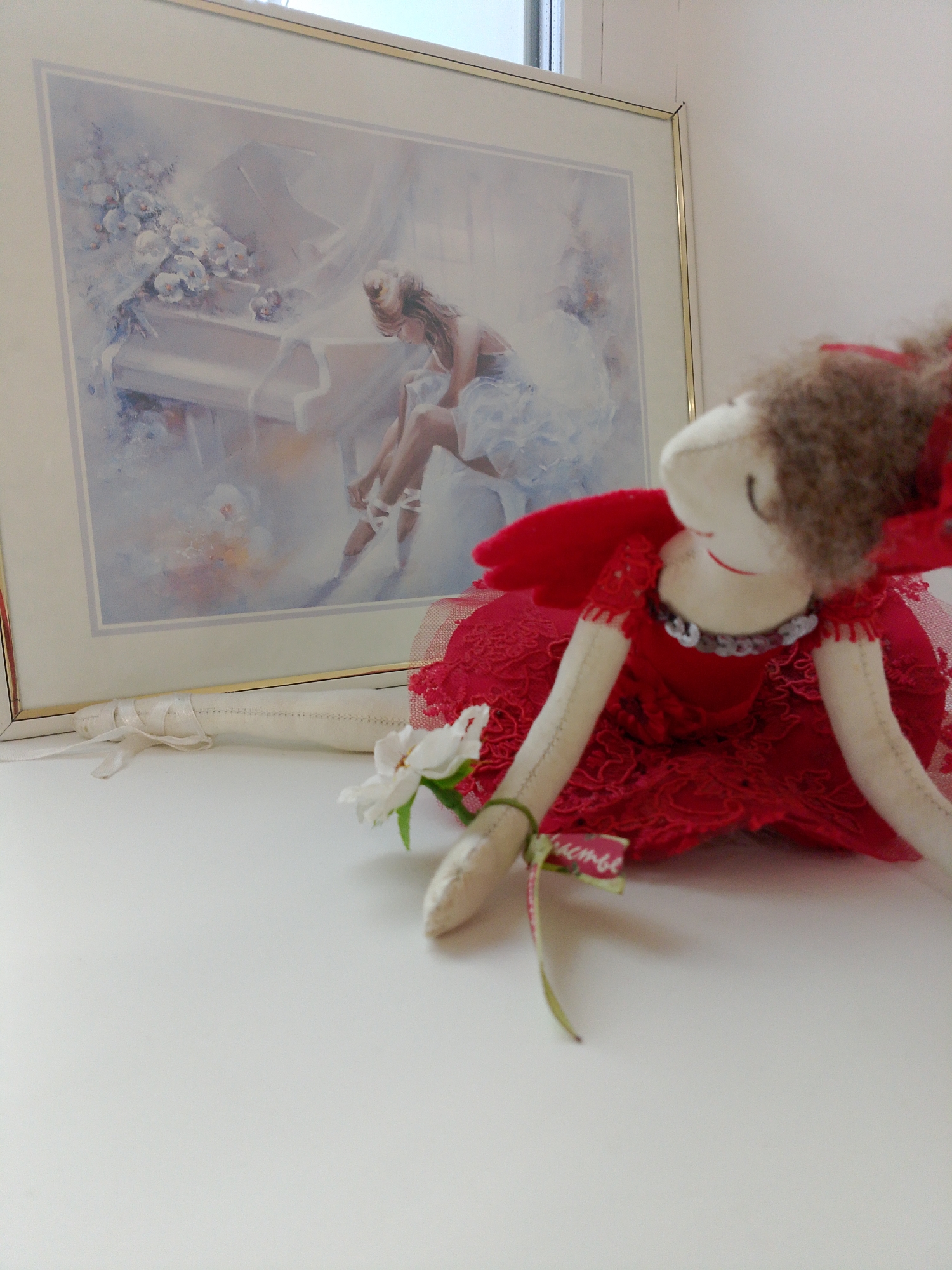 A still life photograph captures a handmade doll in a red dress, positioned on a white surface in front of a framed artwork. The artwork depicts a woman sitting at a white piano in a hazy, impressionistic style. The doll is positioned in a split, with one leg extending towards the left side of the frame and the other tucked beneath her red dress. The doll's right hand holds a white flower. The background is out of focus but appears to be an interior space, possibly near a window, judging by the light. There is no indication in the image that the location is in Izhevsk, Russia. The scene is suggestive of ballet or dance.