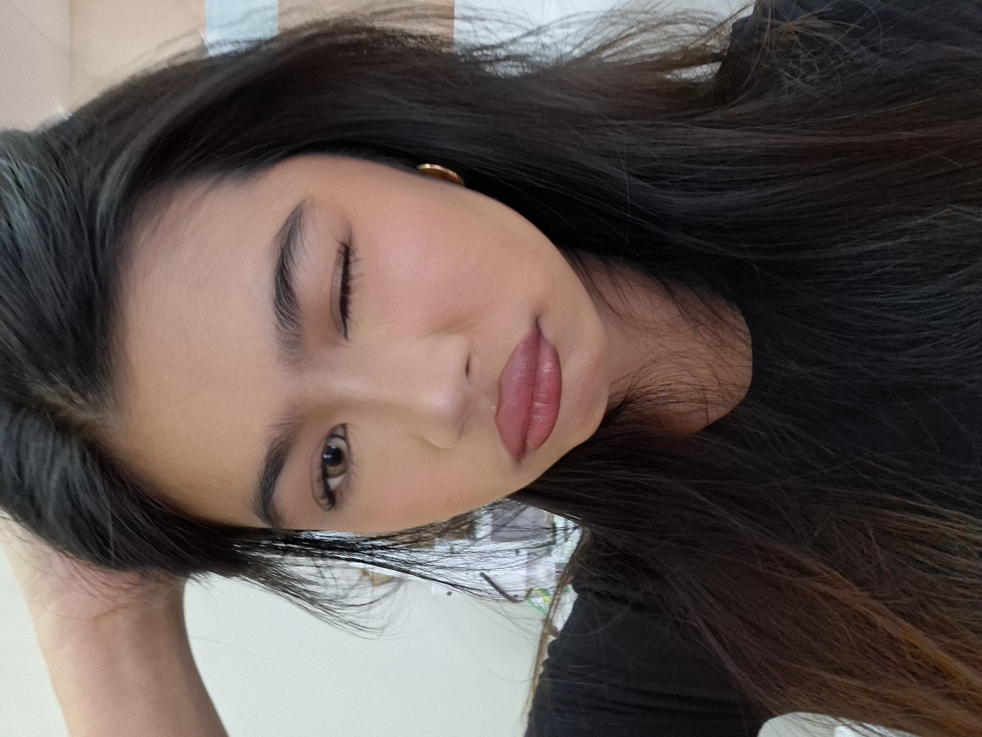 A young woman with long, dark hair, wearing a black shirt and a gold hoop earring, poses for a selfie. She has her left hand on the back of her head and is winking with her left eye. Her makeup includes mascara and lipstick. The background appears to be an indoor setting with a light-colored wall. The image does not provide any information about the location in Kuta, Indonesia.