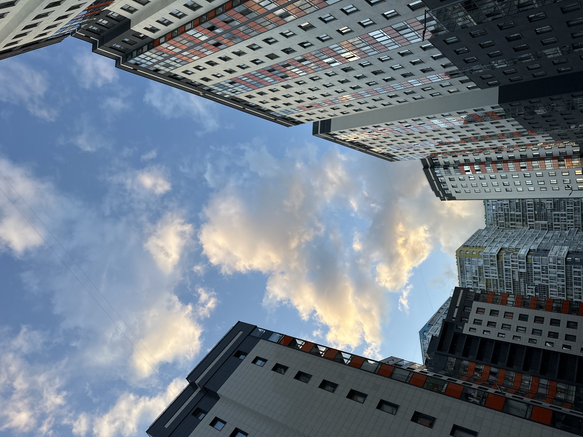 The image depicts a low-angle view of a cluster of modern, multi-story residential buildings in Kudrovo, Russia. The buildings are primarily characterized by their grey facades, interspersed with sections of orange and glass, giving them a contemporary aesthetic. Many windows are visible, reflecting the sky and clouds. The sky above is a mix of blue and white clouds, some of which are illuminated by what appears to be late afternoon or early evening sunlight, casting a warm glow. Two thin, dark wires stretch diagonally across the upper portion of the sky, a common sight in urban environments. The perspective suggests these are newly constructed or recently developed high-rise apartment complexes. No people are visible in the frame. The overall scene conveys a sense of urban development and modern architecture against a natural sky backdrop.