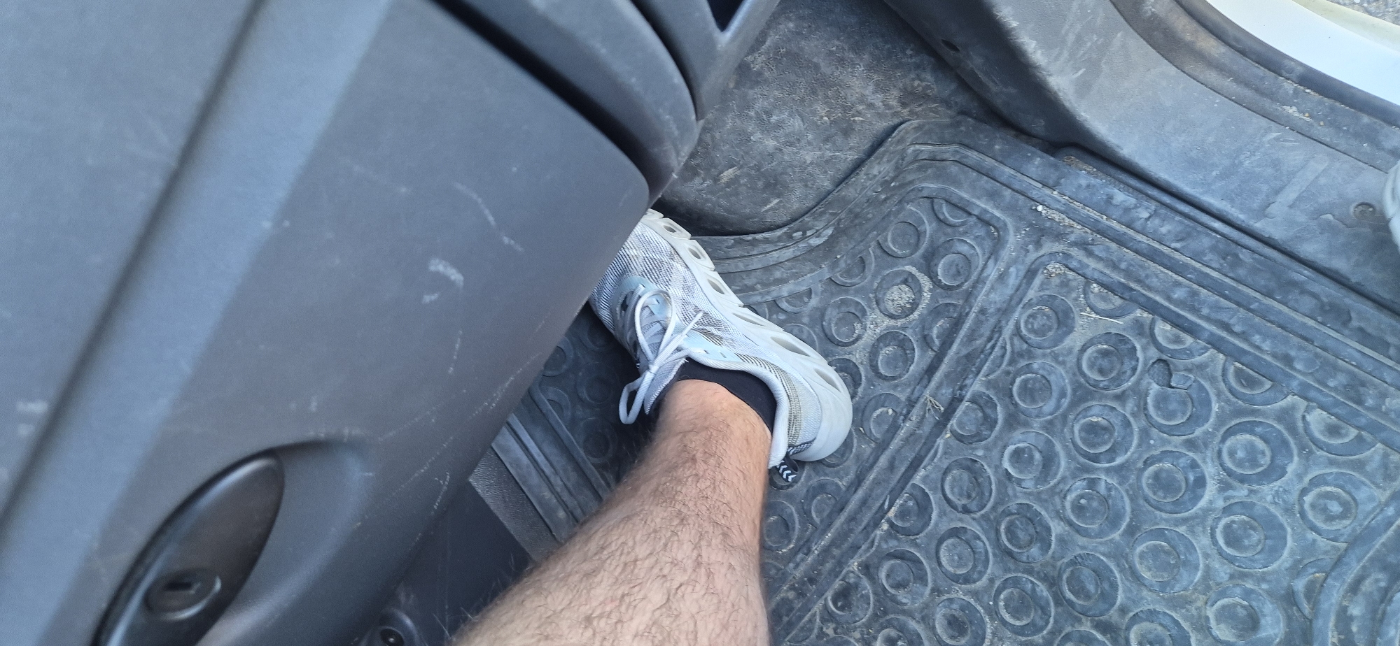 A close-up, eye-level, medium shot captures a person's hairy leg and light gray athletic shoe pressed against a textured black rubber floor mat inside a vehicle. The shoe is positioned on the mat, with the laces visible. The interior of the vehicle appears to be dark gray plastic with some scuff marks. The lighting suggests an outdoor environment or a well-lit interior. The image is centered on the foot and leg, with the surrounding vehicle interior providing context.