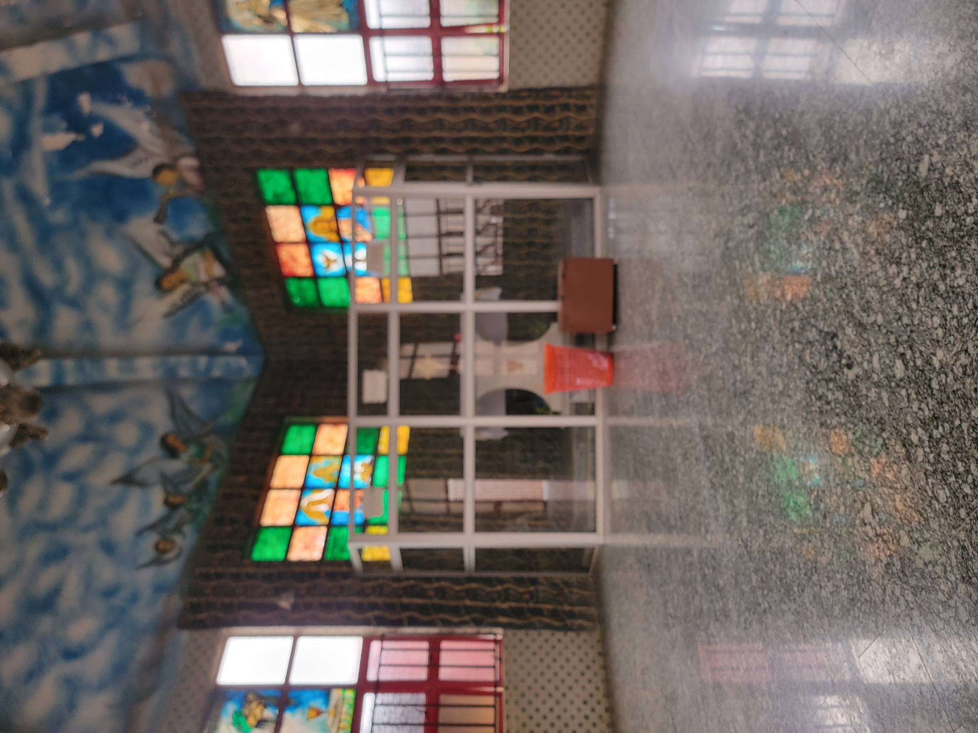 The image depicts the interior of a church or religious building in what appears to be Maiburiji, Nigeria. Stained glass windows are visible on the walls, with a depiction of what looks like an ocean scene with figures on the upper portion of the wall. There is a doorway-like structure with white frames. A small brown table or cabinet with a red plastic bucket is situated near the doorway. The floor is a marbled gray tile.