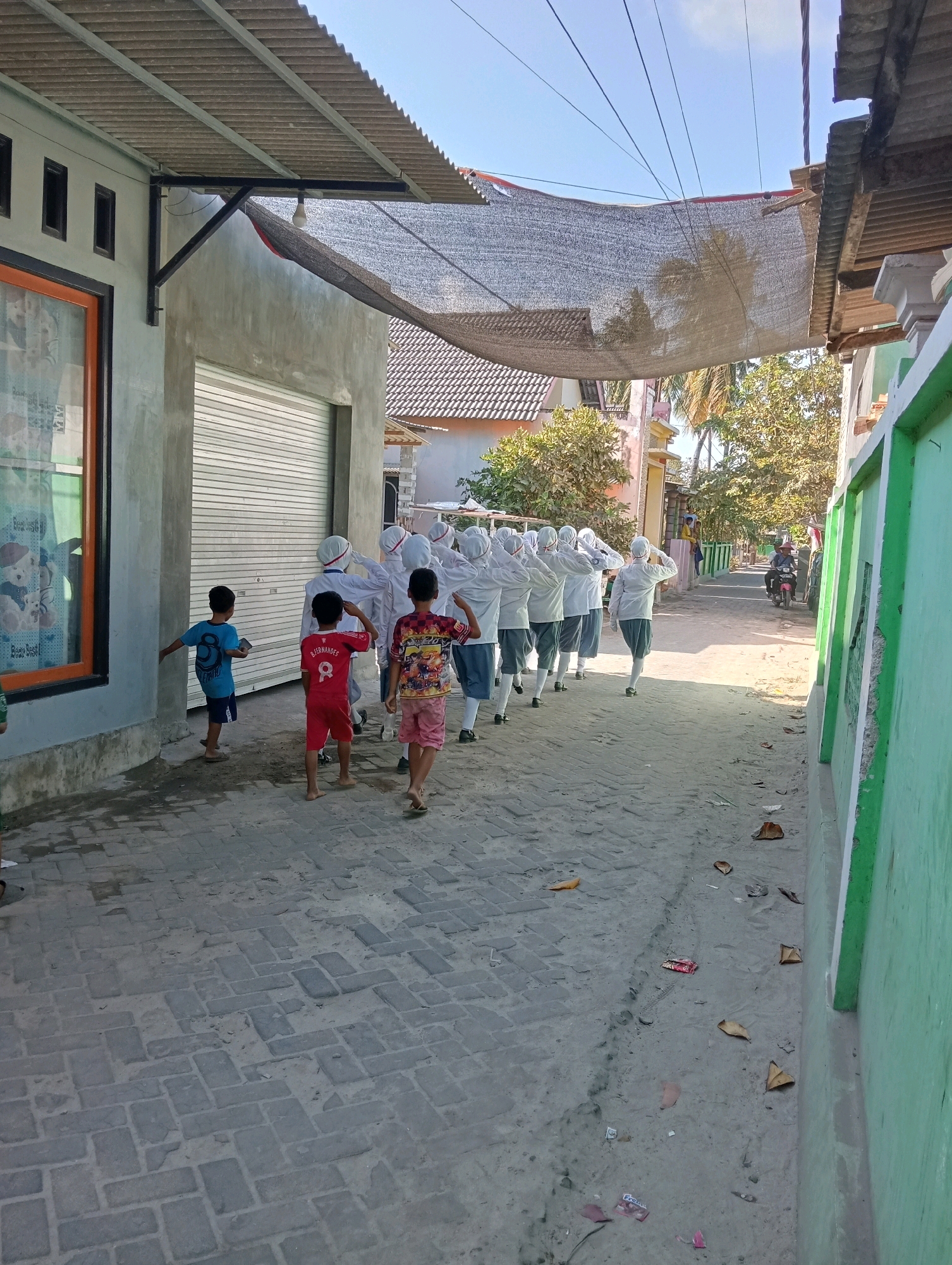 In the city of Pagerungan Kecil, Indonesia, a group of individuals, some dressed in white with head coverings and dark skirts, are walking in a procession down a narrow, paved street. Several children are also present, one in a blue t-shirt and dark shorts, another in a red shirt with the name "B. FERNANDES" and the number 8, and a third in a colorful patterned shirt and pink shorts. They appear to be walking away from the camera, towards a street lined with buildings and vegetation. A motorcycle is visible in the background. The street is covered with interlocking bricks and shows signs of wear and tear. A green wall runs along the right side of the street, and a building with a gray, textured facade and a roll-up door is on the left. A corrugated metal roof covers an awning extending from the building. The sky is clear and blue.