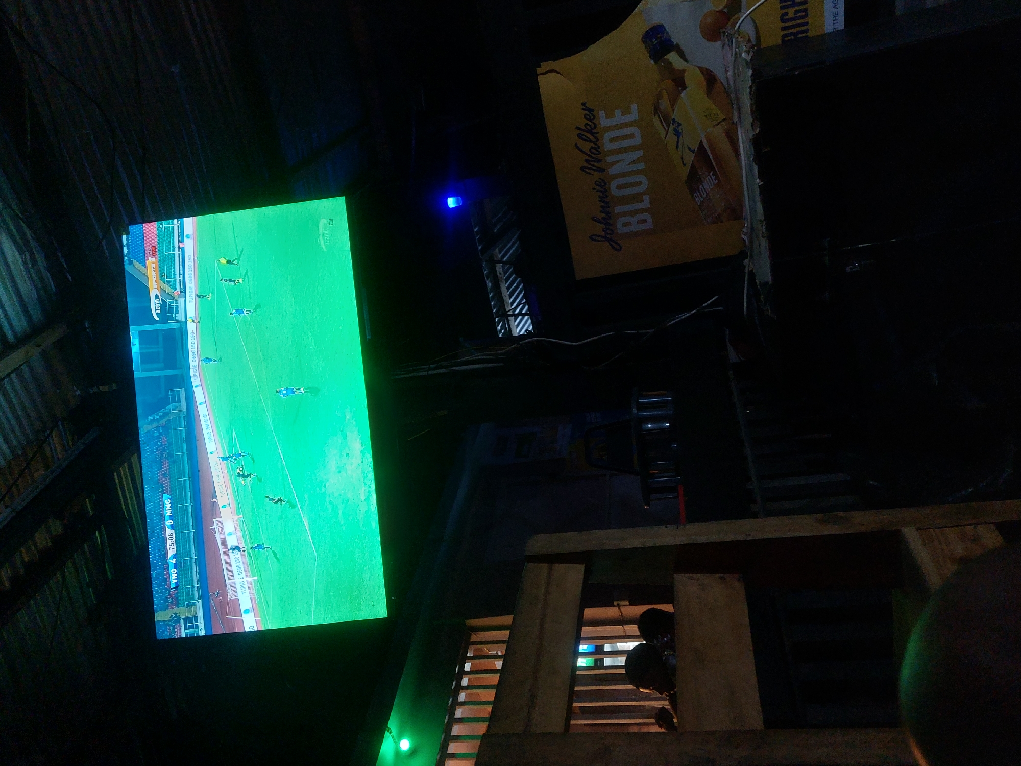 The image captures a dimly lit establishment in Dar es-Salaam, Tanzania, where a football match is being televised. The television screen displays an active game, with the score and team names "YNG" and "MMC" visible. The time on the screen reads "75:08." Advertising information from "TUPIGIE" is displayed on the television screen. In the background, signage for "Johnnie Walker BLONDE" is visible, suggesting the availability of alcoholic beverages. The structure of the building is rough, with corrugated metal roofing. Below the television, a partially visible wooden structure or enclosure can be seen. The overall atmosphere suggests an informal gathering place for watching sports.