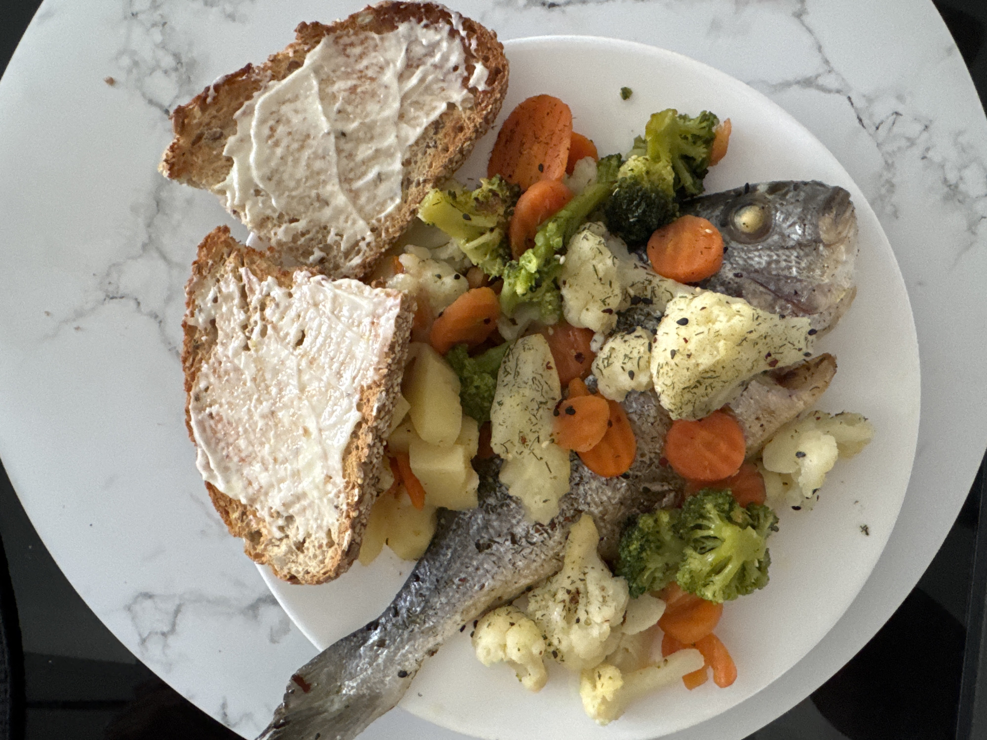 A whole cooked fish, likely sea bream, is presented on a white plate with a marble-patterned placemat underneath. The fish is arranged with its head to the right, and its tail extending to the left. It is surrounded by an assortment of cooked vegetables, including broccoli florets, sliced carrots, and cauliflower florets. Diced potatoes are also visible beneath the fish. Two slices of toasted bread, generously spread with a white substance, possibly mayonnaise or cream cheese, are placed on top of the vegetables near the fish's head. The scene is illuminated by overhead lighting, casting subtle shadows on the food and plate. The overall setting appears to be a casual meal, possibly in a dining area. There are no people visible in the image, and no specific indicators of the location being in Finestrat, Spain.