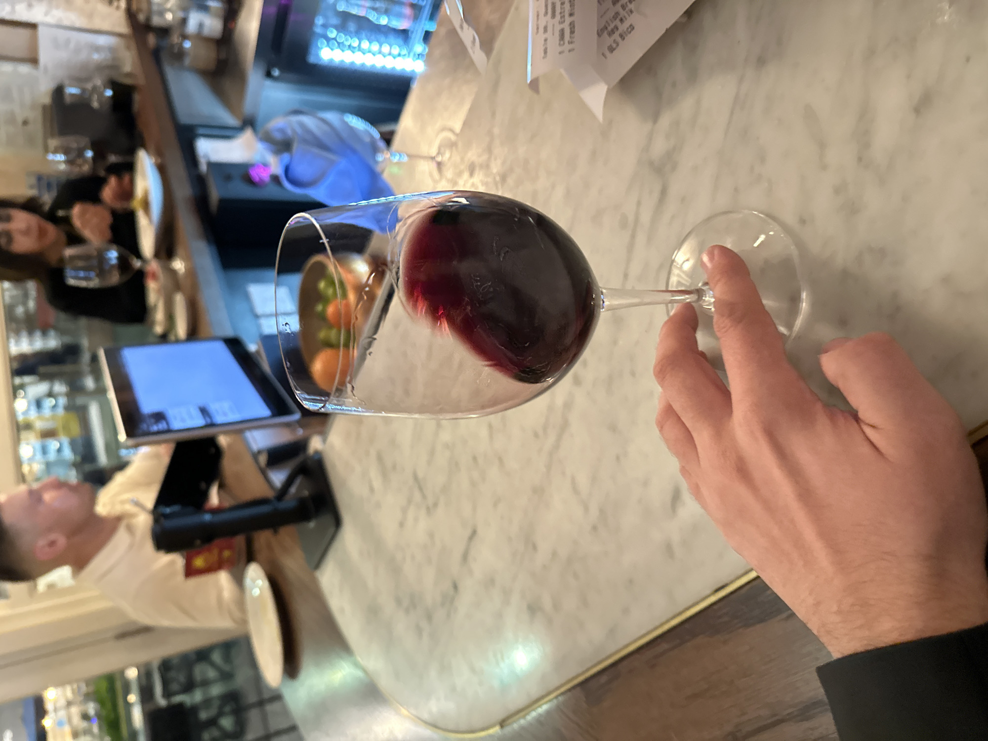 The image displays an indoor scene within a bar or restaurant located in the City of London, United Kingdom. In the immediate foreground, a fair-skinned hand, partially visible on the left and wearing a dark sleeve, holds a wine glass containing red wine that is actively being swirled, evidenced by the motion blur of the liquid. The hand and glass rest on a white marble countertop with grey veining and a brass-colored edge trim. To the right of the glass, a crumpled white paper receipt is visible, displaying partial text including "Table No." and menu items.
Behind the marble counter, a dark wooden bar surface extends, where two individuals are seated. On the left, a light-skinned male with short hair, wearing a light-colored polo shirt, is focused on a tablet with a blue screen mounted on a black stand. An empty white plate and a red cup are situated near him. To the right, a blurred dark-haired female in a dark top appears to be consuming food from a white plate; a wine glass is also present near her. A bowl containing citrus fruits (oranges and limes) is visible on the wooden bar behind the foreground wine glass. Further in the background, a beverage cooler with blue interior lighting is partially visible on the right.