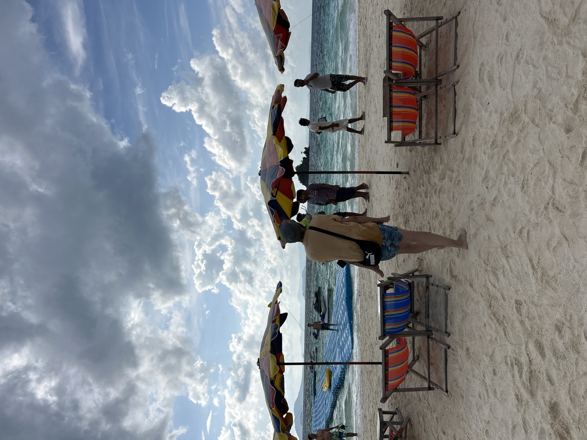 The image depicts a tropical beach scene in Phru Nai, Thailand. The foreground is dominated by sandy beach with several colorful striped lounge chairs and two large, multi-colored beach umbrellas. In the middle ground, a blue floating platform with a yellow kayak is visible in the turquoise ocean water. Several people are scattered across the beach and on the floating platform, some walking, others standing. Jet skis are visible in the water behind the platform. The background shows a hazy coastline with mountains and a clear blue sky filled with dramatic clouds. The overall atmosphere is one of leisure and outdoor activity.