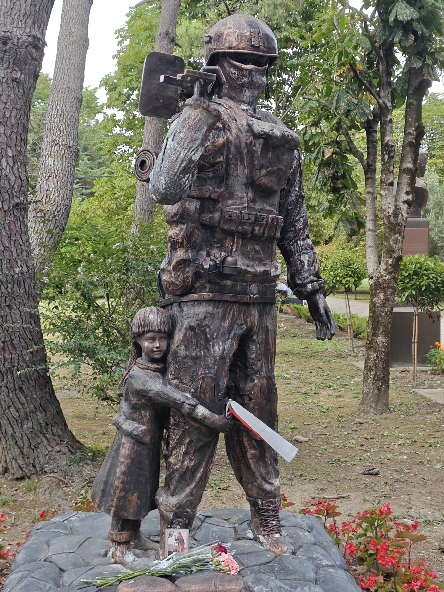 This is a full shot of a bronze sculpture in a park-like setting, depicting a soldier and a child. The soldier stands tall, wearing a helmet, tactical vest, and holding a weapon over his shoulder. He appears to be looking forward. A young child, dressed in a coat, is embracing the soldier's leg. The sculpture is placed on a dark, rocky base. In front of the base, there are flowers and a small religious icon. The background consists of trees, green foliage, and a pathway. The overall scene suggests a memorial or a tribute.