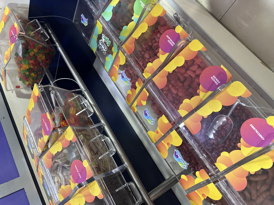 The image displays a detailed view of a bulk candy dispensing station within a retail establishment. Two tiers of clear acrylic dispensers are visible. The upper tier, mounted on a metallic rail against a dark blue background, holds various gummy candies, including elongated multicolored sour strips labeled "TIRA ÁCIDA," two-color gummy snake candies labeled "VIBORA TWO COLORS," and a bulk quantity of multicolored gummy bears. The lower tier consists of multiple "Dulcipolis" branded clear acrylic dispensers, featuring orange and yellow circular patterns on their sides, resting on a white counter. These dispensers contain a variety of red-toned candies: dark red bean-shaped candies labeled "CHECOLINES," with a metal scoop immersed in them; bright red irregular-shaped candies labeled "GOMITA SANDIA"; and red, chili-dusted mango-flavored candies labeled "MANGO ENCHILADO." Another dispenser on the lower right displays a "Snak Club Tajín" logo. No individuals are present in the scene. The overall environment is clean and brightly lit, characteristic of a commercial candy store or a dedicated candy section within a larger retail outlet. Given the prominent "Dulcipolis" branding and Spanish labeling, the location is a retail store in Mexico, plausibly one of the numerous Dulcipolis locations found throughout Mexico City.