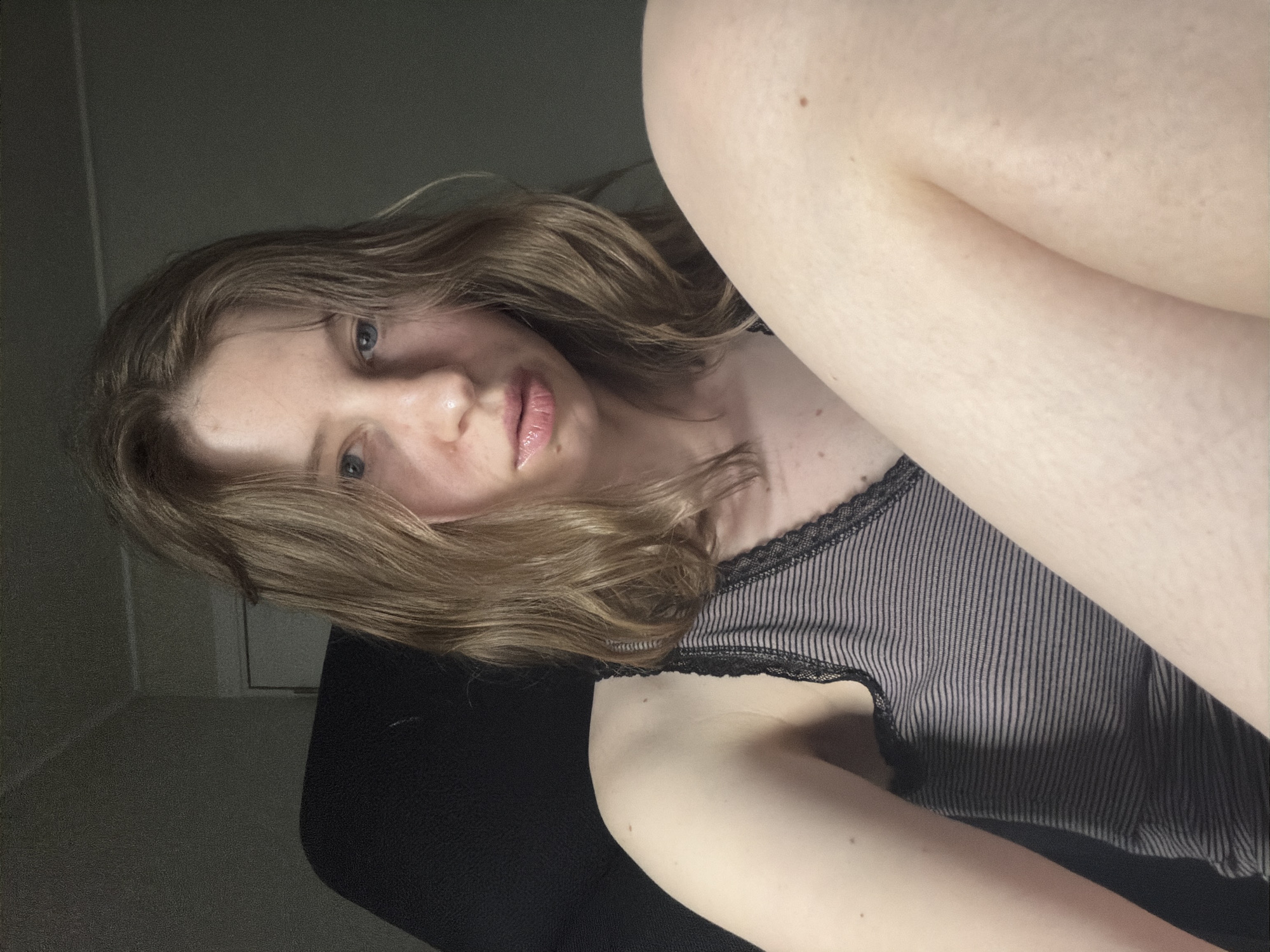 A young Caucasian woman with shoulder-length light brown hair is captured in a vertical, close-up selfie. She is lying down, with her head tilted back and her gaze directed towards the camera. Her eyes are blue, and she has full lips with a glossy sheen. She is wearing a black and grey striped tank top with lace trim. Her skin is pale, with a few freckles visible on her arm and shoulder. The lighting is dim, suggesting it might be nighttime or a dimly lit room. The background is mostly dark, with a hint of a wall and possibly furniture. The woman appears relaxed, with a neutral expression. The image was taken in Dongducheon-si, South Korea. There is no visible text in the image.