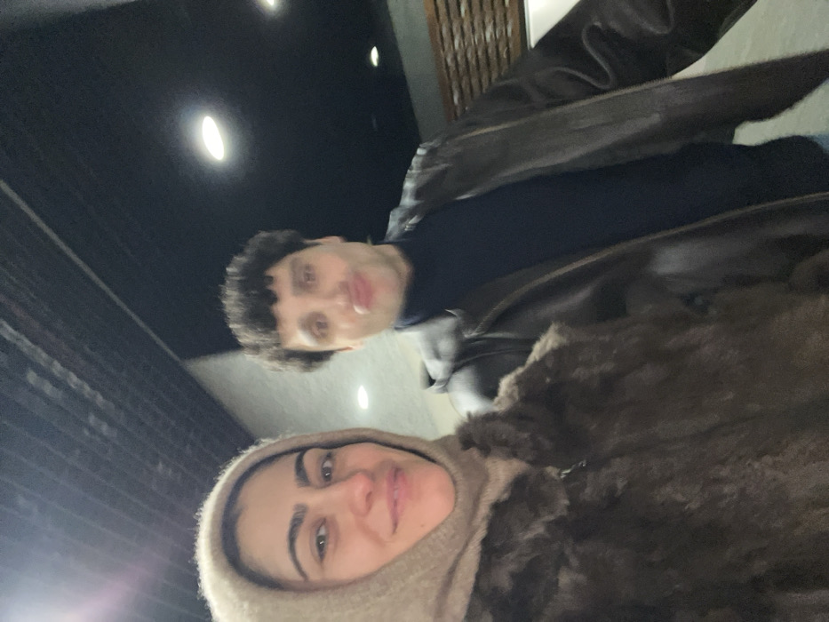 A selfie is taken at an upward angle, showcasing two individuals. A woman with dark hair and fair skin wears a beige hood-like garment. Above her and slightly out of focus is a man with dark, short hair, a dark sweater, and a brown leather jacket. The background consists of a dimly lit setting with visible architectural elements and ceiling lights.