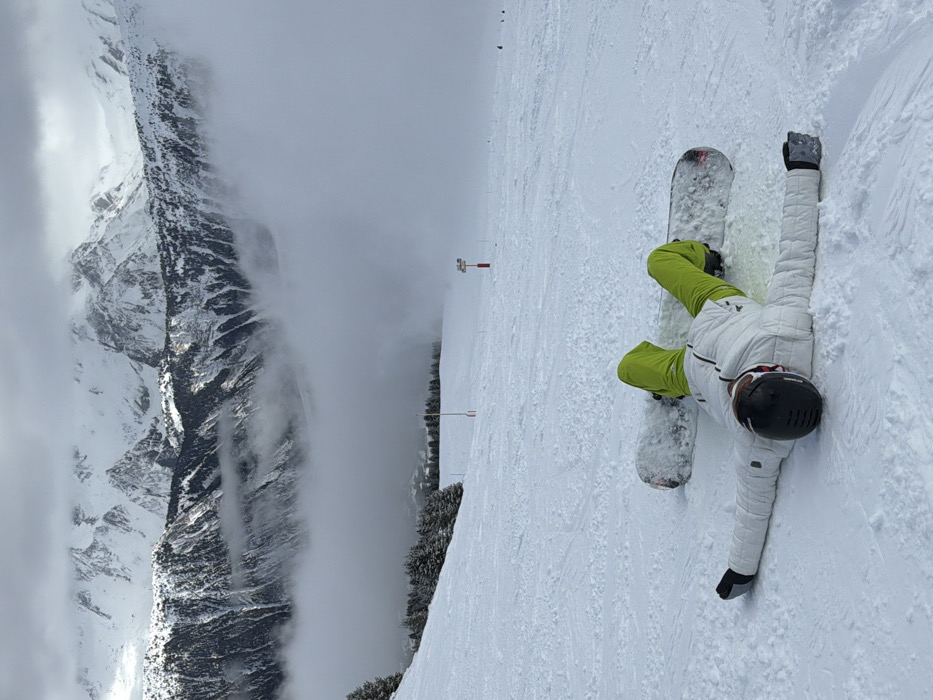 A person lies supine on a snow-covered ski slope in Courchevel, France. The individual is wearing a white puffer jacket, bright lime green ski pants, a black helmet with a red stripe, and black gloves. Their arms are extended outwards, and a snowboard is attached to their feet, resting flat on the snow beside them. The person's head is oriented towards the top of the image. The foreground features groomed snow. To the left, a line of snow-dusted pine trees borders the slope. In the mid-ground, a village with multiple buildings is visible through a break in the low-lying clouds or fog. Two red-tipped ski marker poles are present further up the slope. The background is dominated by a majestic, snow-capped mountain range, partially obscured by heavy clouds or fog, indicating an overcast sky.