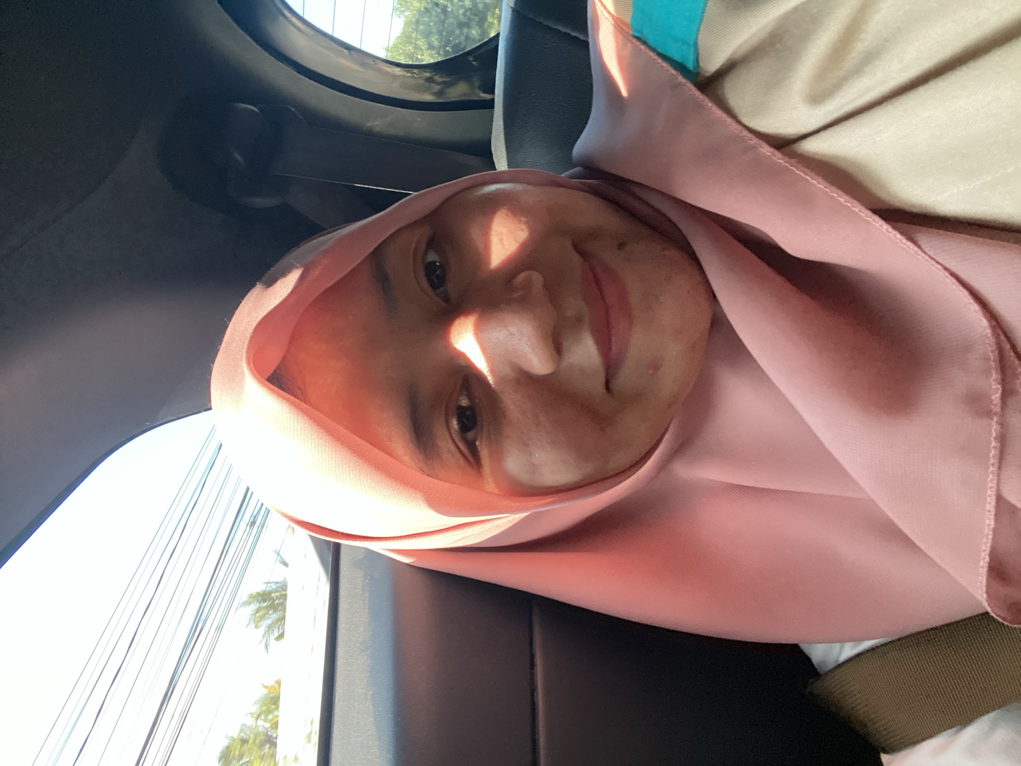 A young woman, likely of Indonesian descent, is photographed from the chest up, inside a vehicle. She is wearing a light pink hijab that covers her hair and neck, with a portion of her face illuminated by a ray of sunlight. She has warm skin tone and is smiling faintly, with her eyes looking towards the camera. Her attire also includes a beige jacket with a teal accent. The background shows the interior of a car, with a dark grey seat and headliner, and the window revealing a bright, overcast sky with visible power lines and some greenery, suggesting an outdoor environment within the city of Astambul Kota, Indonesia.