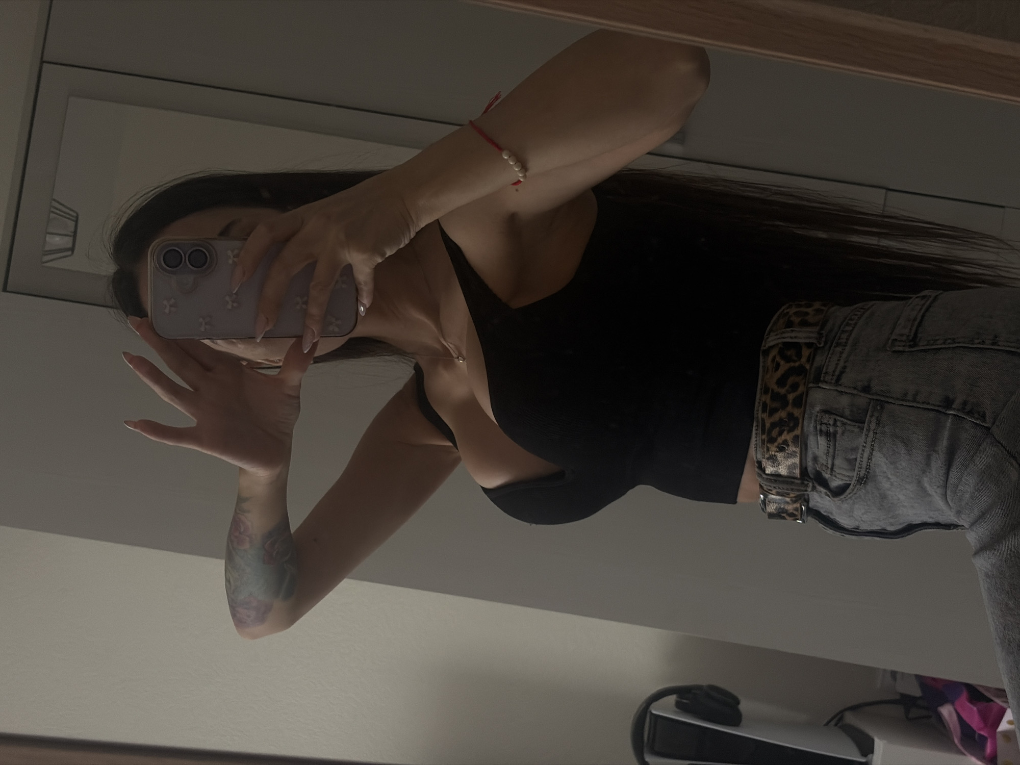 A woman is taking a selfie in a mirror. She is wearing a black top and grey jeans with a leopard print belt. She has long, dark hair. A tattoo is visible on her left arm. The phone she is using has a lavender case with white flowers. In the background, a white object with a black accessory and a pink and white object are visible. The location is not identifiable as Saint Petersburg, Russia.