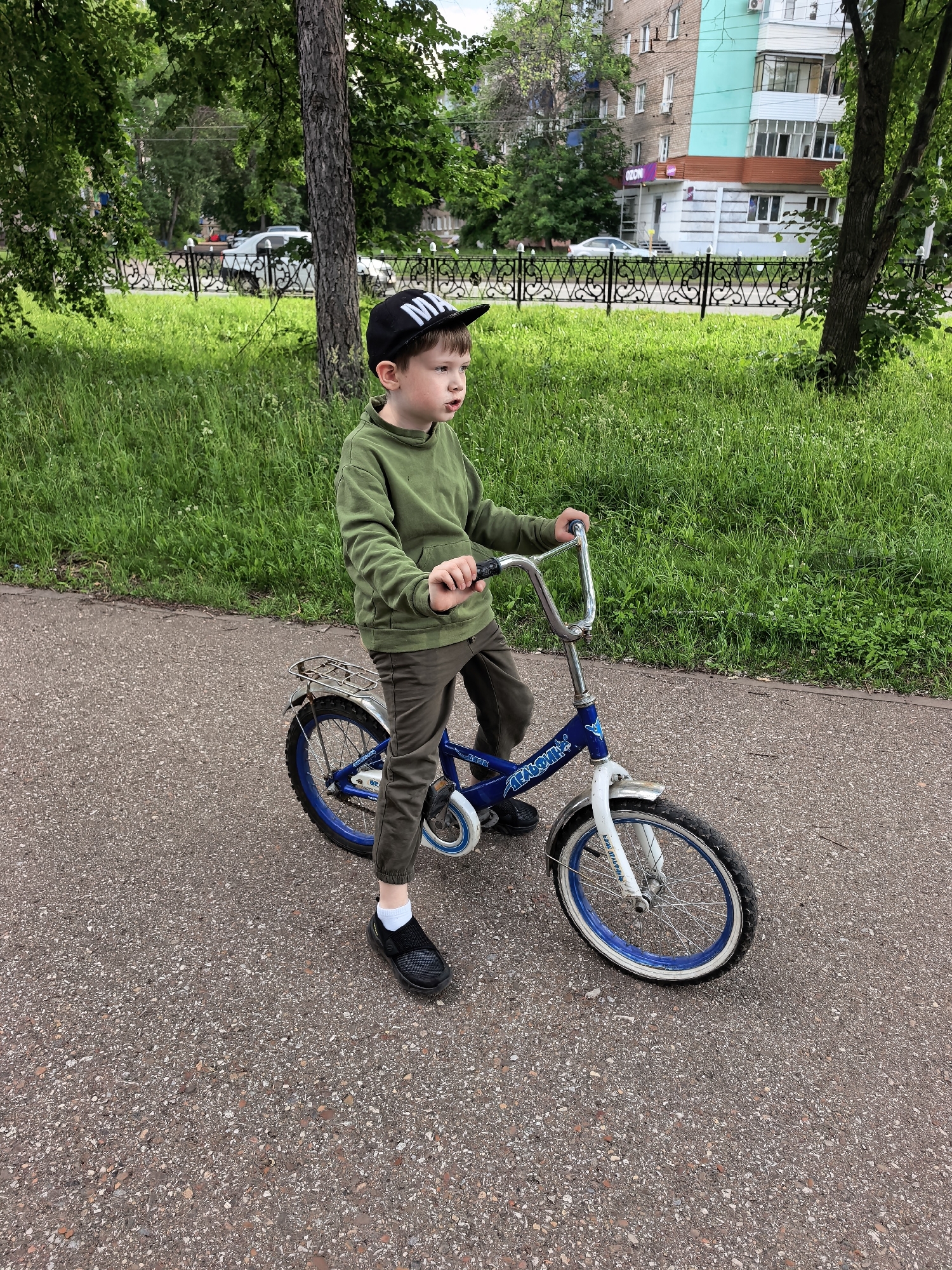 This image depicts a young boy and his bicycle on an asphalt path in a park-like setting within the city of Sterlitamak, Russia.
The central figure is a young boy, approximately 5-7 years old, with short brown hair, wearing a black baseball cap with partially visible white lettering "M^" on the front. He is dressed in a dark olive green hooded sweatshirt, dark olive green or brown trousers, white ankle socks, and black slip-on athletic shoes. His gaze is directed towards the right side of the frame, and his mouth is slightly ajar. He stands to the left of a child's bicycle, gripping the chrome handlebars with both hands. His left foot is planted on the asphalt path, while his right foot is elevated near the bicycle's pedal.
The bicycle is blue with white accents. It features a chrome rear rack, a white front fender, and blue wheel rims with white sidewall tires. The word "МЕЧТА" (Mechta, Russian for "Dream") is visible in white lettering on the bicycle's frame.
The ground beneath the boy and bicycle is an asphalt path composed of dark grey aggregate. To the left and directly behind the path is a lush area of green grass. Further back, a decorative black metal fence with repeating circular and diamond patterns separates the grassy area from a paved street. Several vehicles, including a white SUV and a silver sedan, are visible on the street.
In the background, beyond the street and fence, are multi-story residential or commercial buildings. One prominent building exhibits a light blue (teal) section on its upper facade and a section with white and grey striped walls at the ground level, topped by a reddish-brown awning. A purple and white "OZON" sign is mounted on the facade of the ground-level establishment, indicating a Russian e-commerce pickup point. Multiple windows, balconies, and external air conditioning units are visible on the buildings. A large tree with dark bark and green foliage stands prominently in the mid-ground, partially obscuring the buildings. The sky is overcast, suggesting a daytime scene with diffuse lighting.