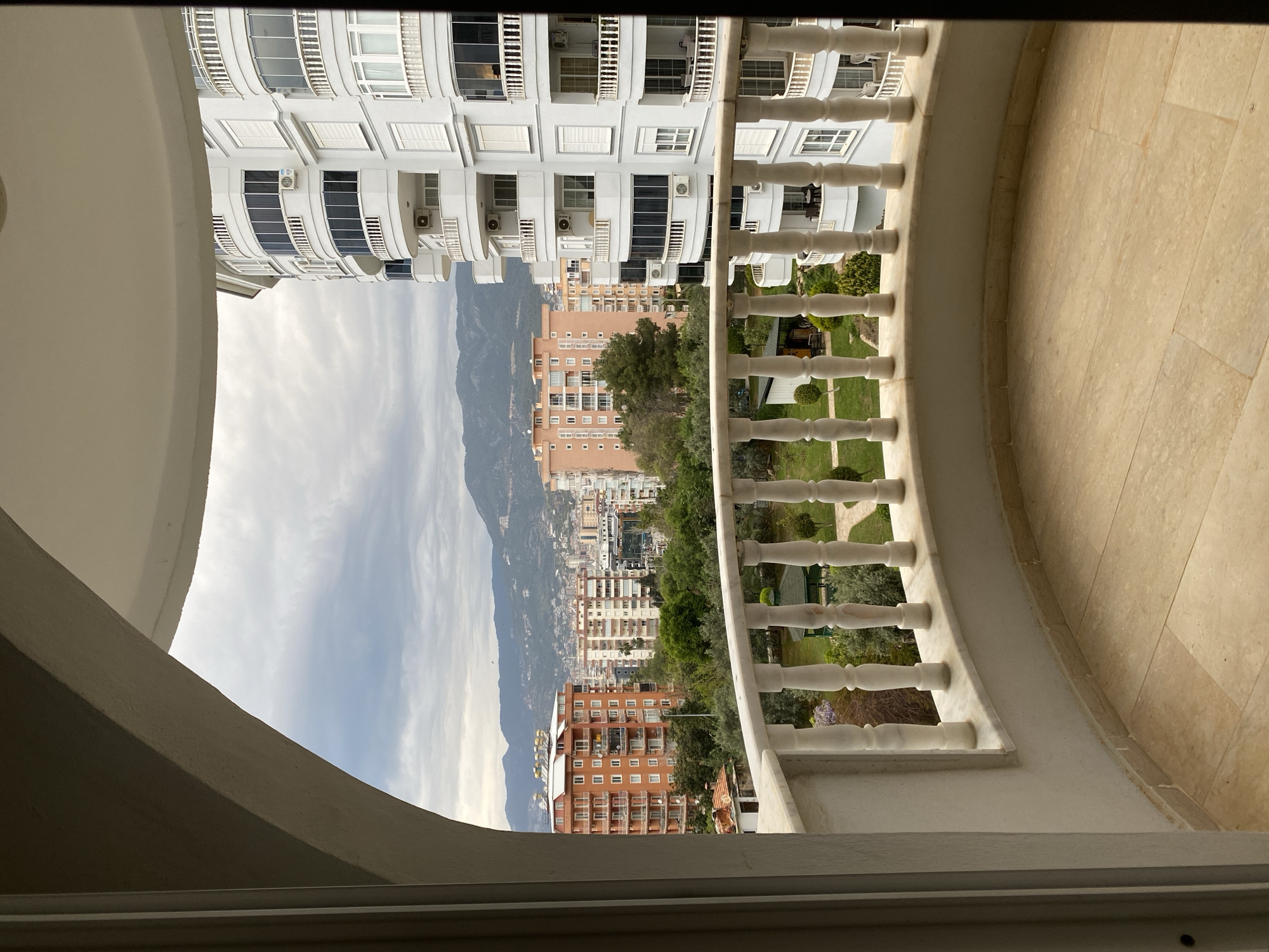 The image presents a view from a balcony, presumably in Alanya, Turkey, given the context. The balcony features a white, curved balustrade with classic baluster designs, and a tan tiled floor. The viewpoint offers a panorama of the city, revealing an array of buildings including both high-rise white apartment complexes and lower, earth-toned residential buildings. Beyond the buildings lies a landscape highlighted by tree cover and a mountain range in the distance, partially obscured by clouds. The sky is overcast. The photo is taken from within a building, indicated by the window frame at the bottom.
