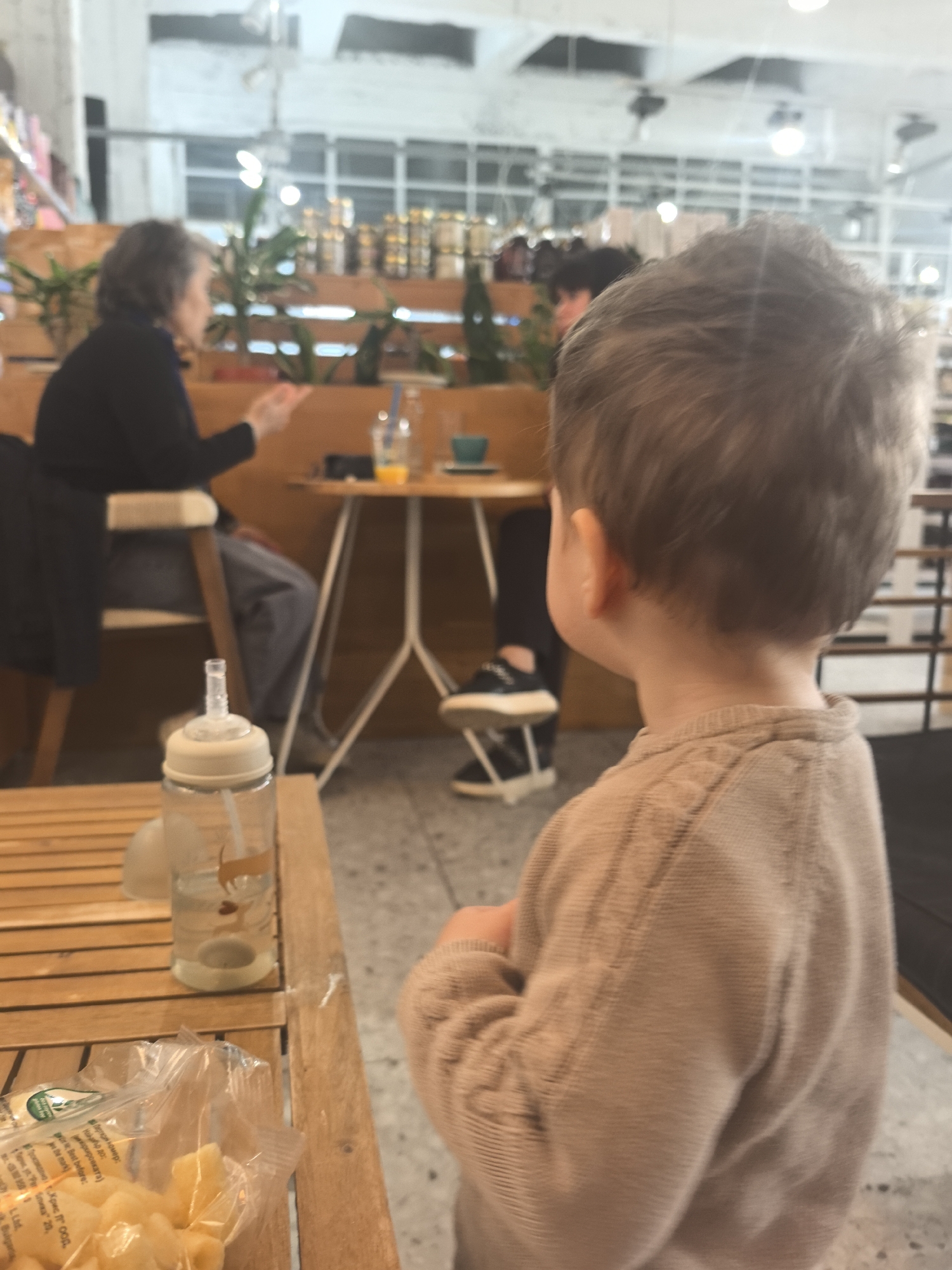 In a cafe in Stara Zagora, Bulgaria, a toddler with light brown hair stands facing away from the camera, wearing a tan-colored sweater. A wooden table is in the foreground. On it are a baby bottle filled with water and a package of snacks. In the background, two people are seated at a small round table with beverages on it, engaged in conversation. The cafe has a bright, airy atmosphere with large windows and greenery visible in the background.